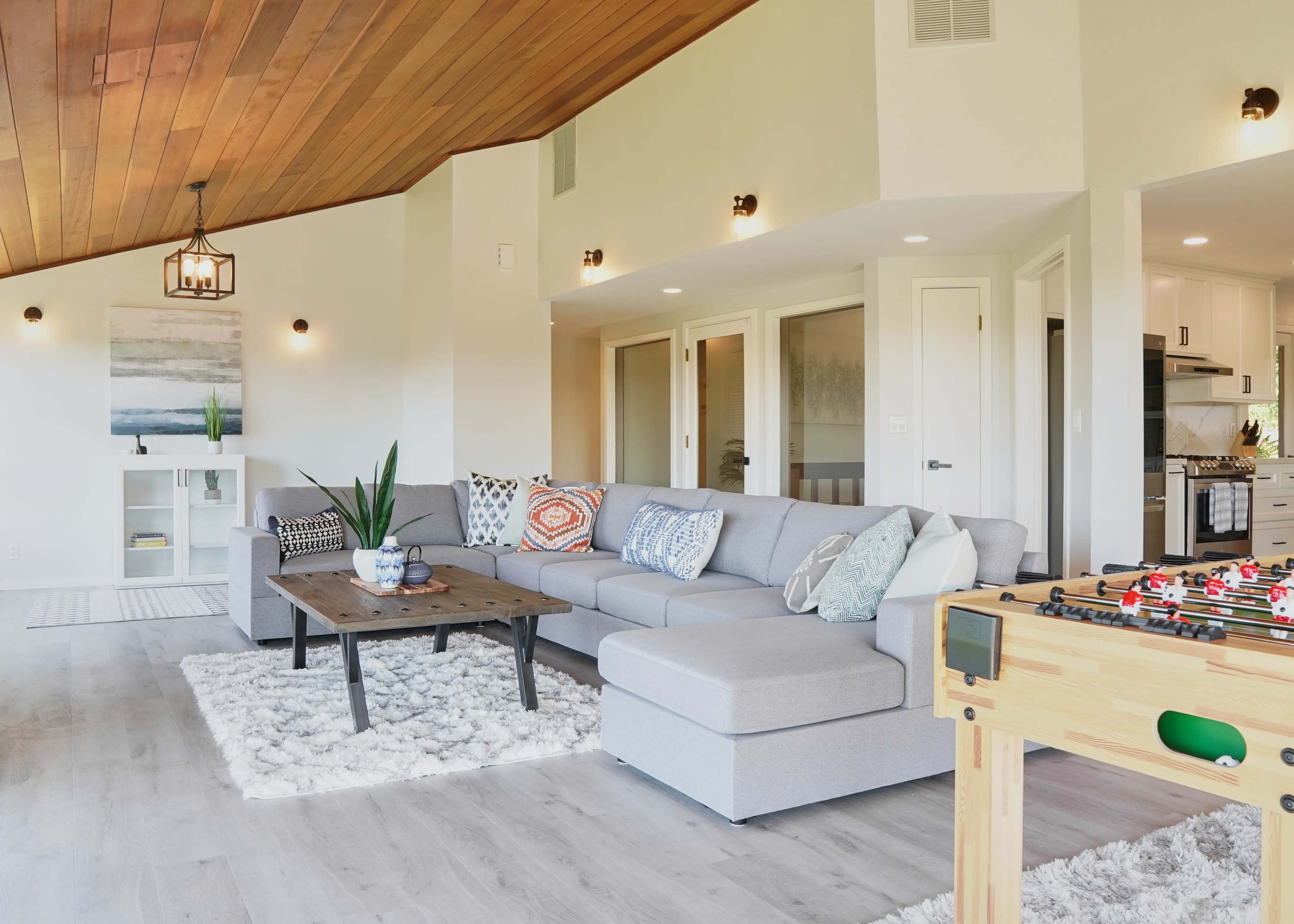 The image shows a modern living room with a gray sectional sofa, a wooden coffee table, and a foosball table in a bright space with a sloped ceiling and decor elements.