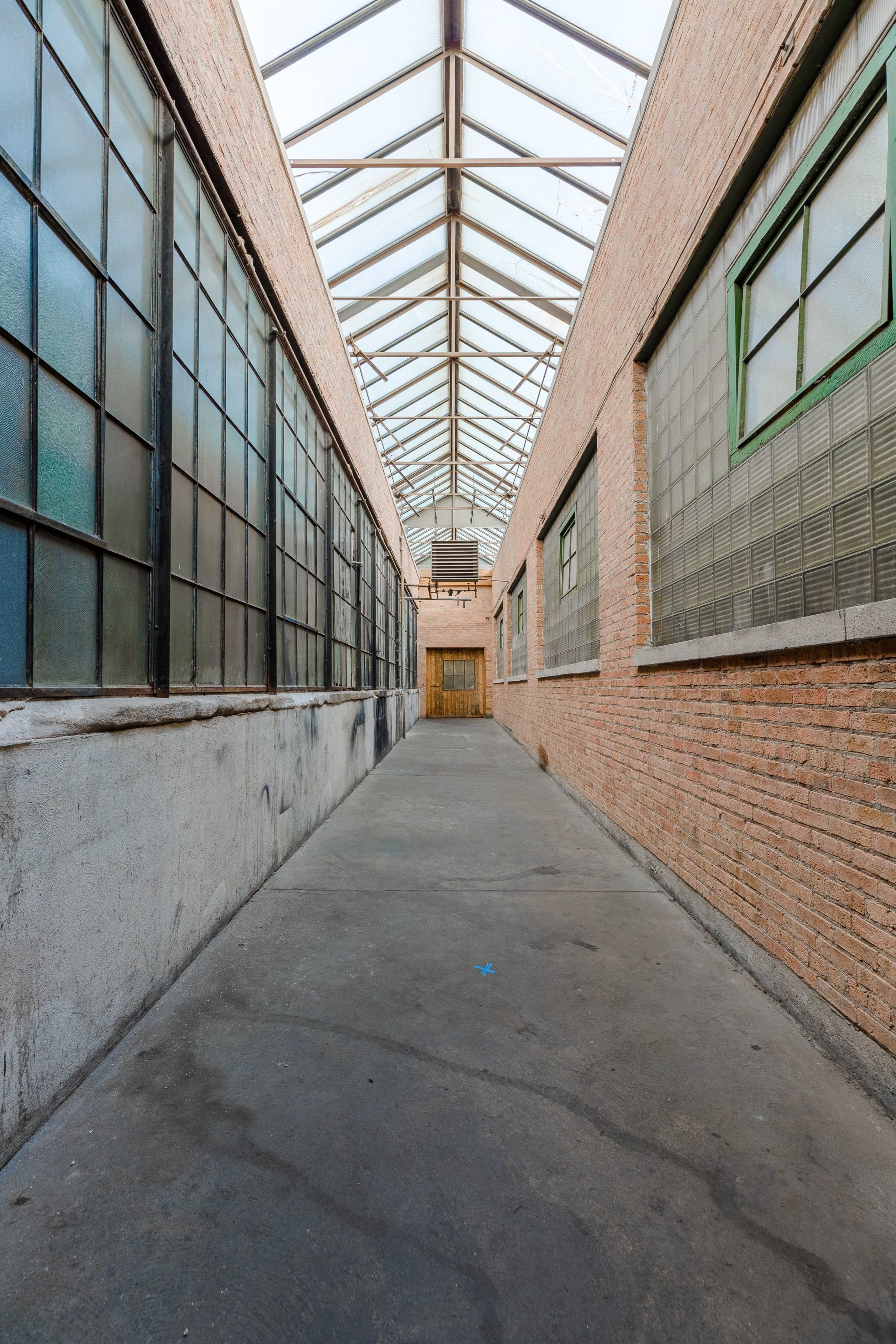A long, narrow corridor lined with brick walls and large windows, leading to a wooden door at the end.