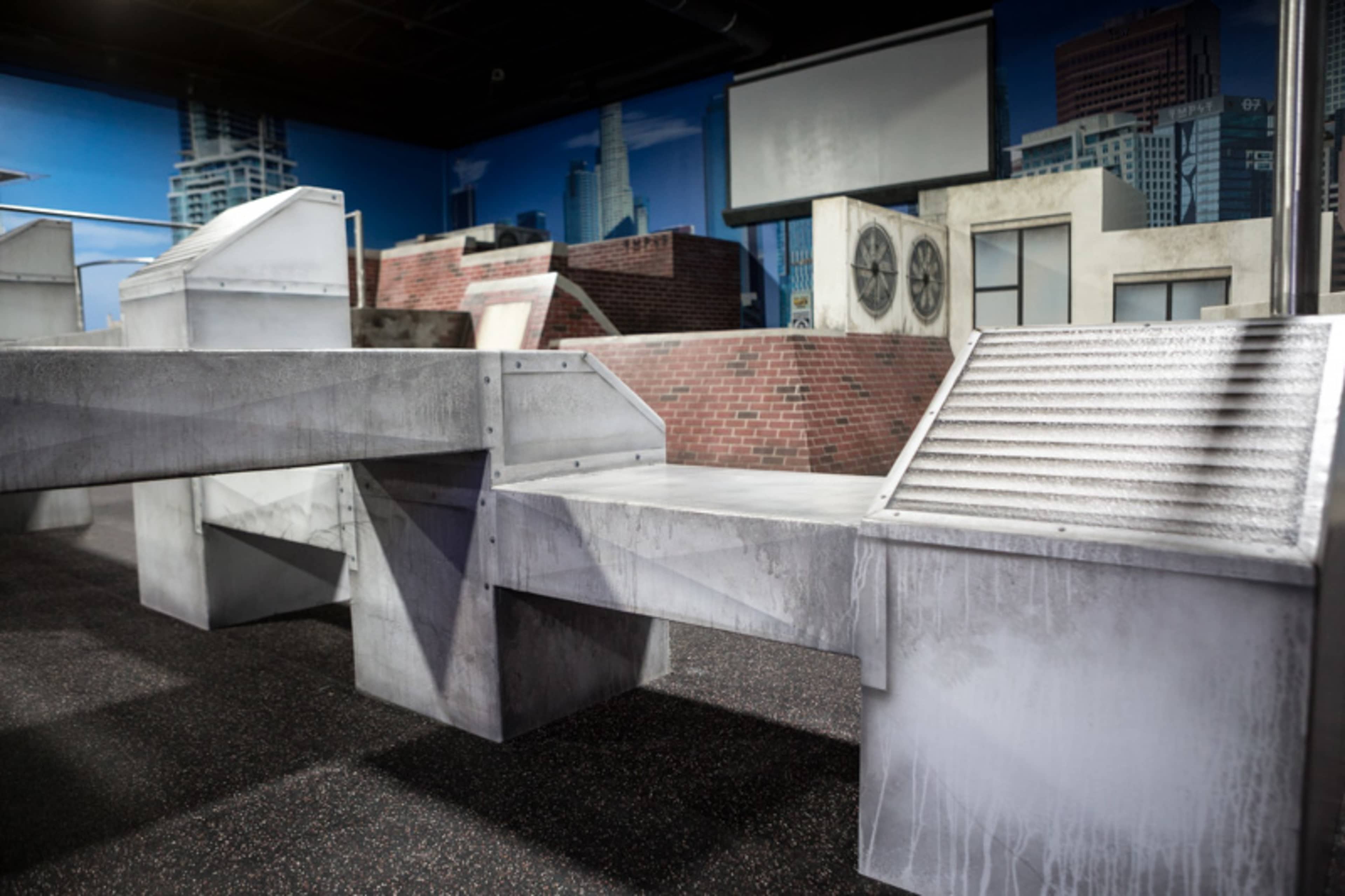 The image shows a gray urban-themed obstacle course with various structures resembling rooftops and ventilation systems against a city backdrop.