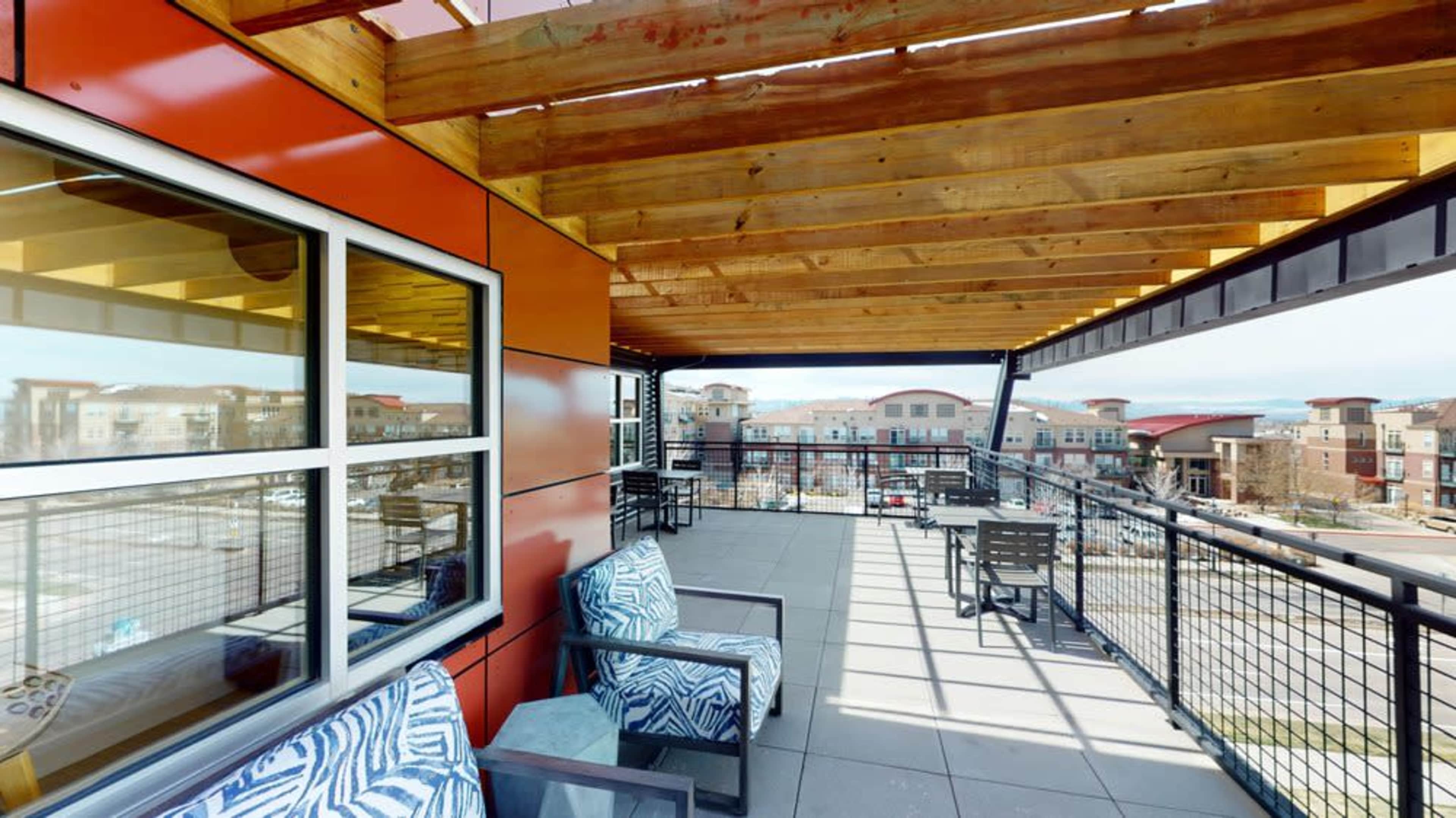 A rooftop terrace with wooden beams overhead, featuring seating areas and a view of nearby buildings.