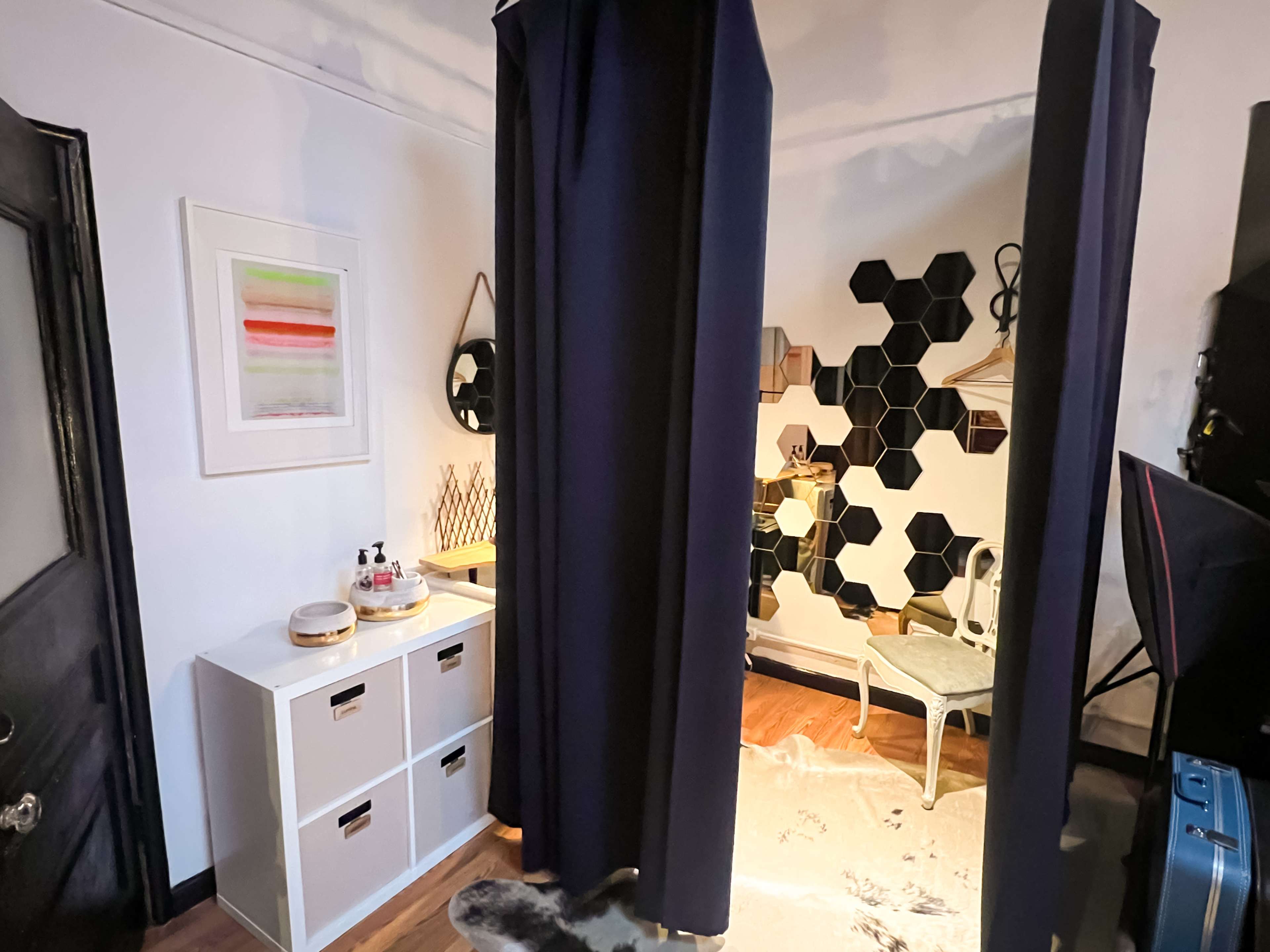 A small room divided by dark curtains, featuring a white storage unit, hexagonal wall decor, and a chair beside a rug.