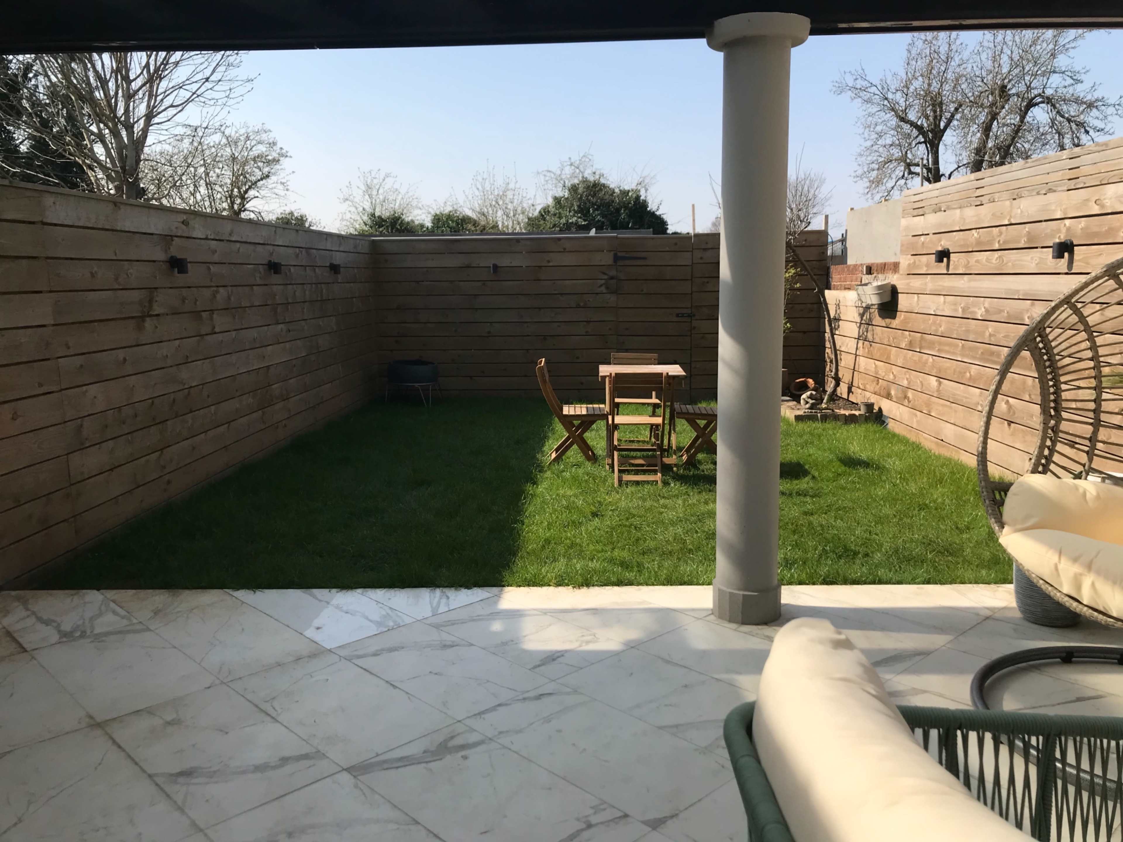 The image shows a small enclosed yard with wooden fence panels, a green lawn, a table with chairs, and a seating area featuring a round hanging chair.