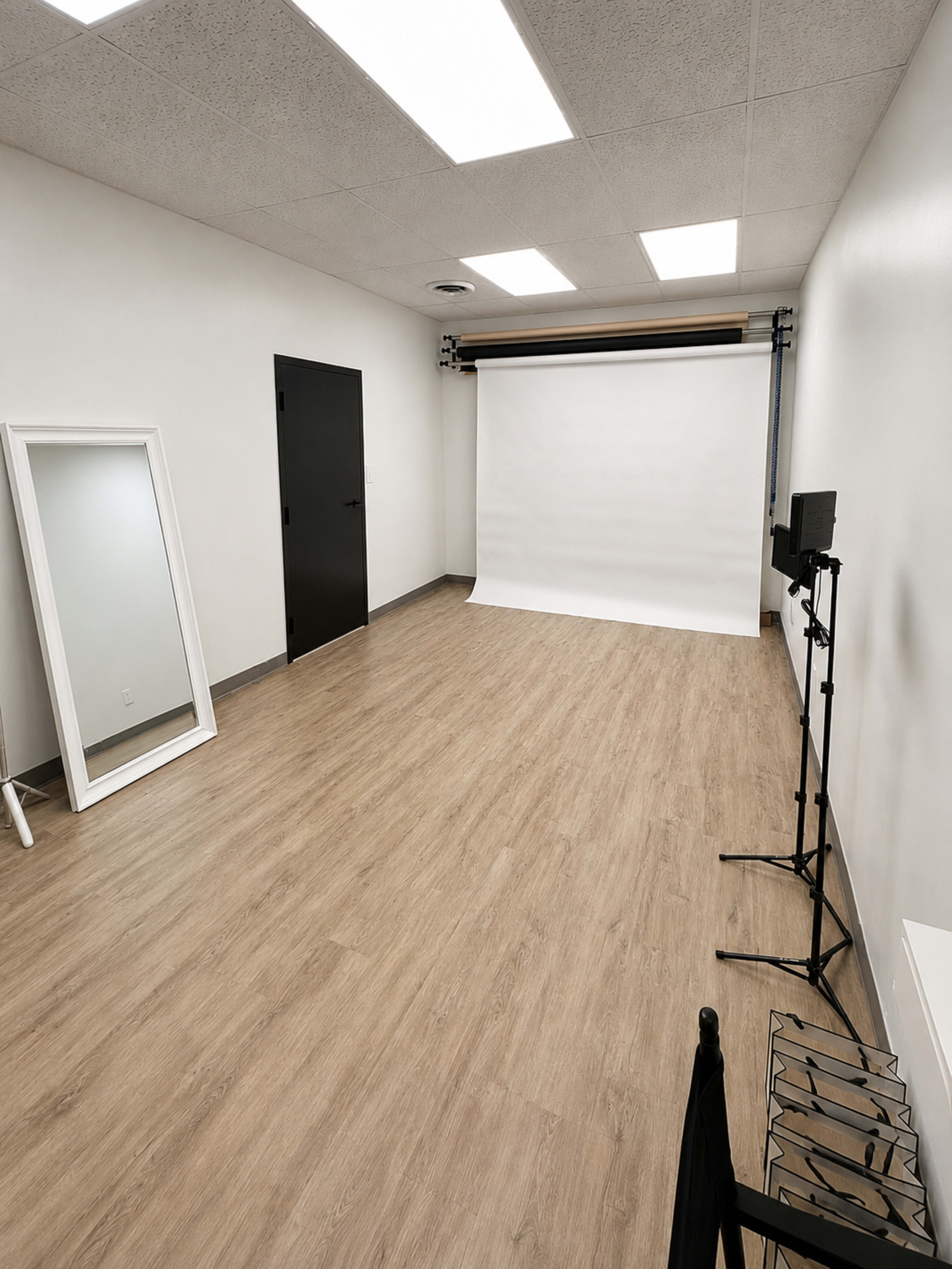 The image shows a minimalistic studio space with a wood-like floor, a white backdrop, a mirror, a black door, and a light stand.