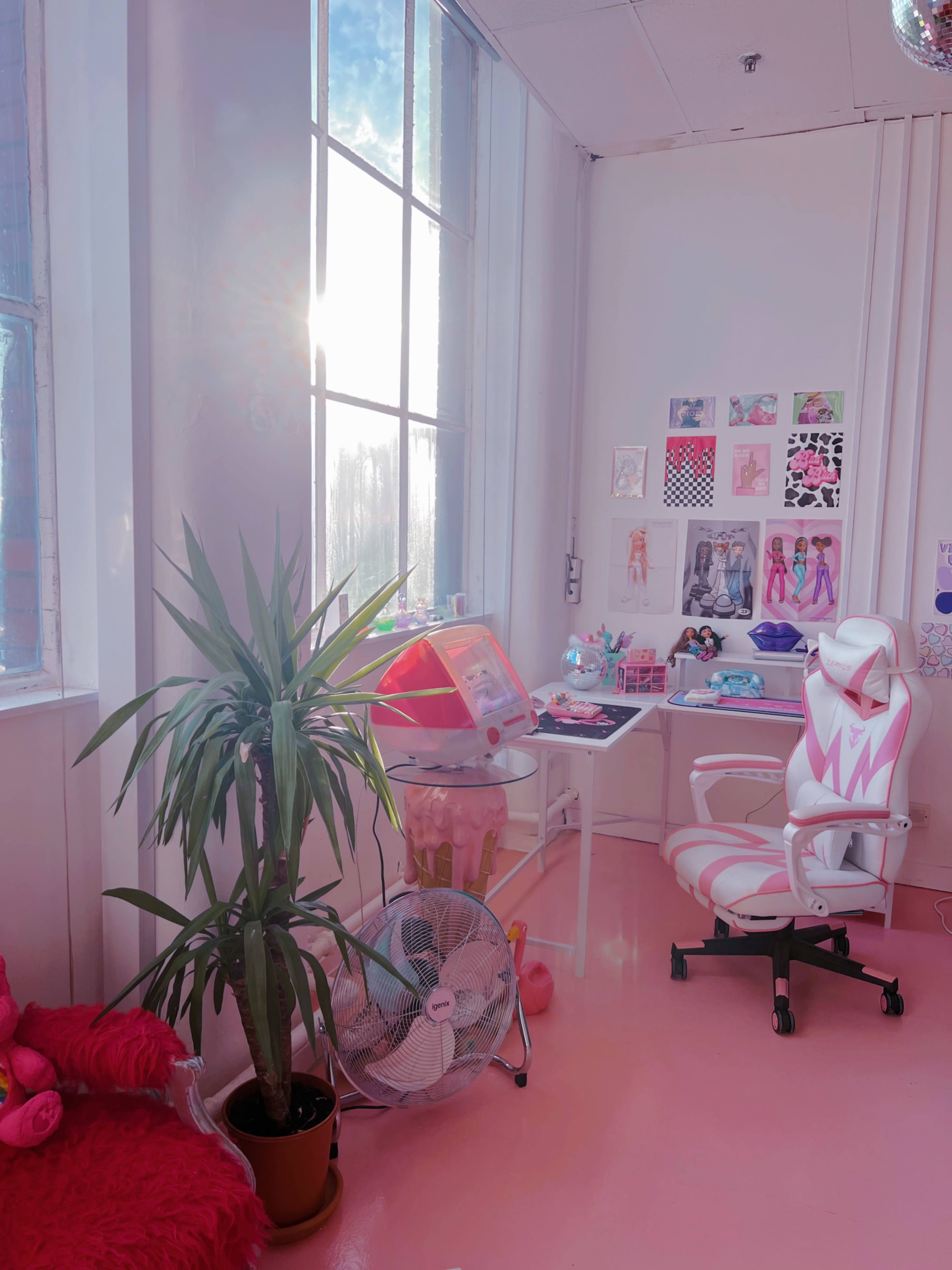 A brightly colored room features large windows, a pink floor, a white desk with various decorations, and a plant beside a fan.