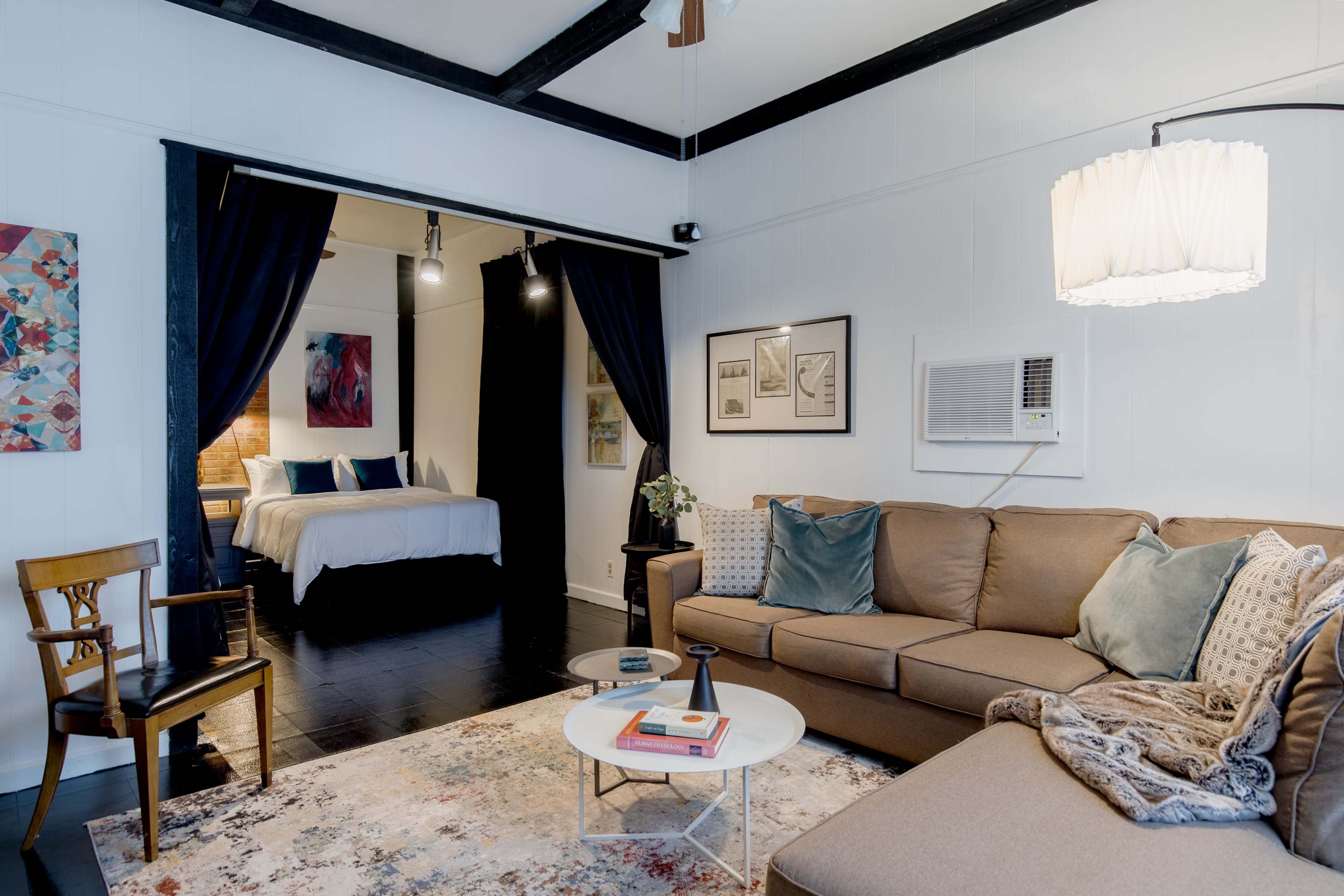 The image shows a living area with a couch, coffee table, and a chair, alongside an open doorway leading to a bedroom with a bed.