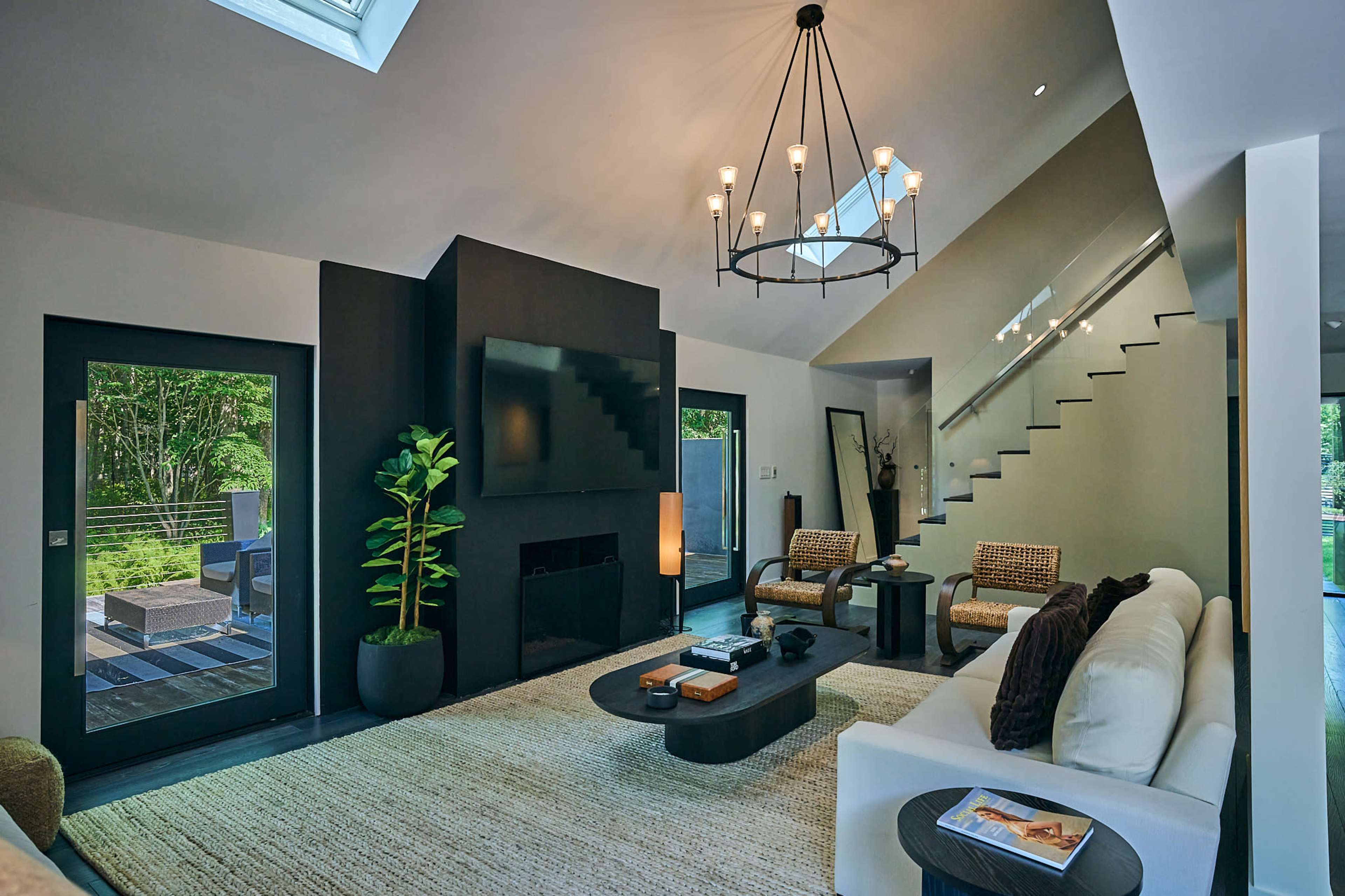 The image shows a modern living room with a minimalist design, featuring a large sofa, two armchairs, a circular coffee table, and a staircase leading to an upper level.