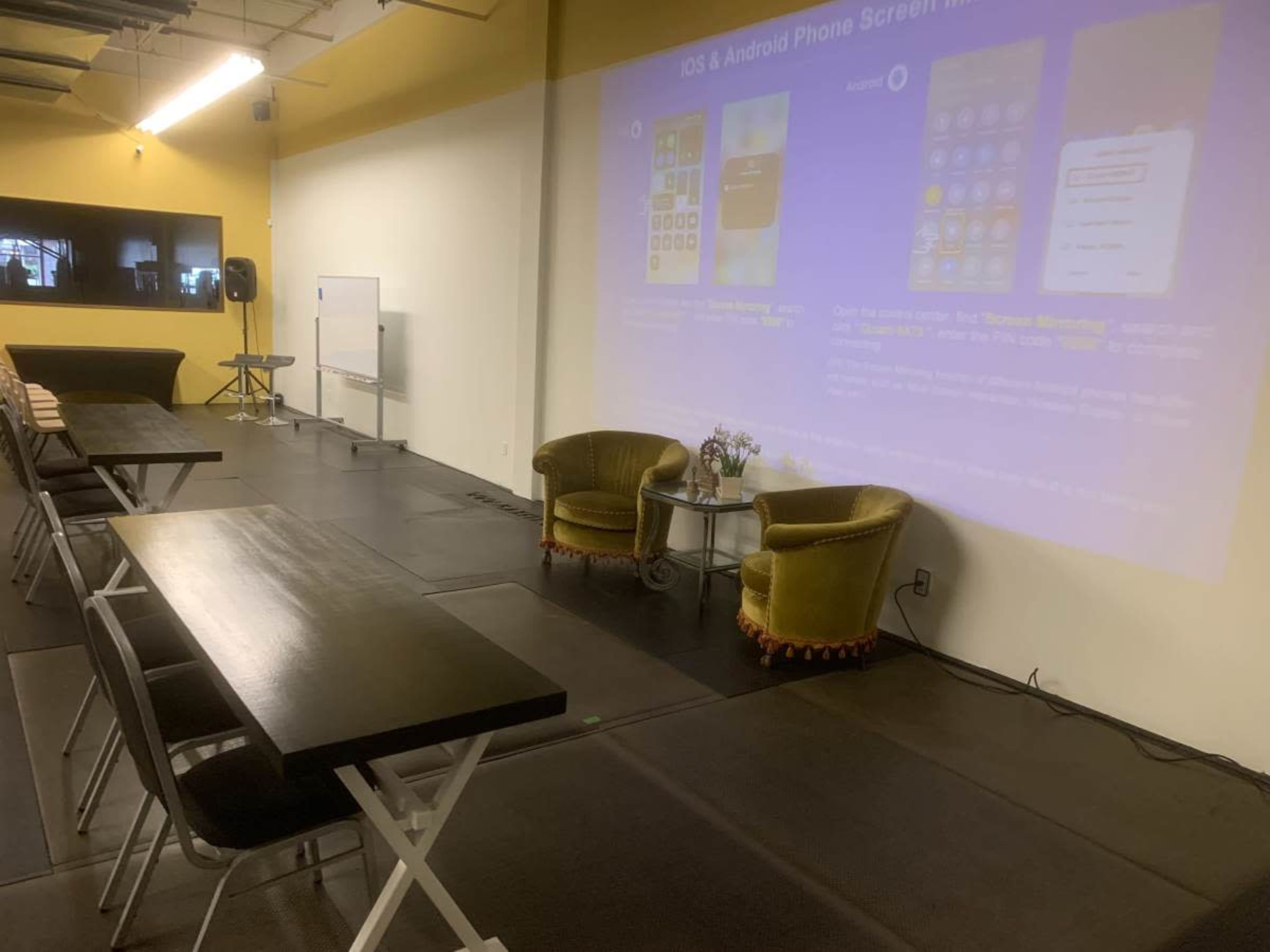 The image shows a room set up for a presentation, featuring two chairs and tables arranged in rows, with a large screen displaying information about mobile phone screens.