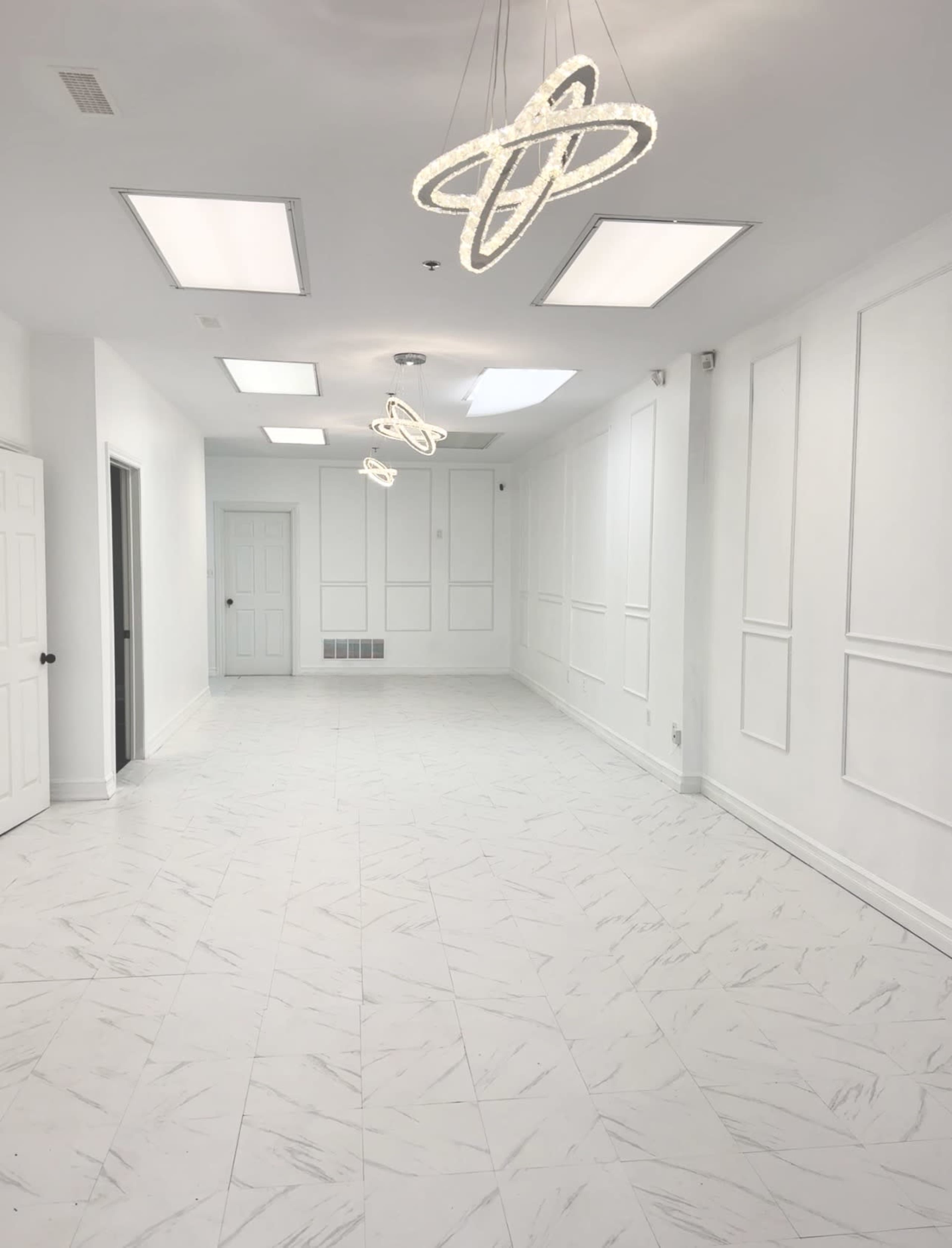 The image shows a spacious, brightly lit interior with white walls, a tiled floor, and modern light fixtures hanging from the ceiling.