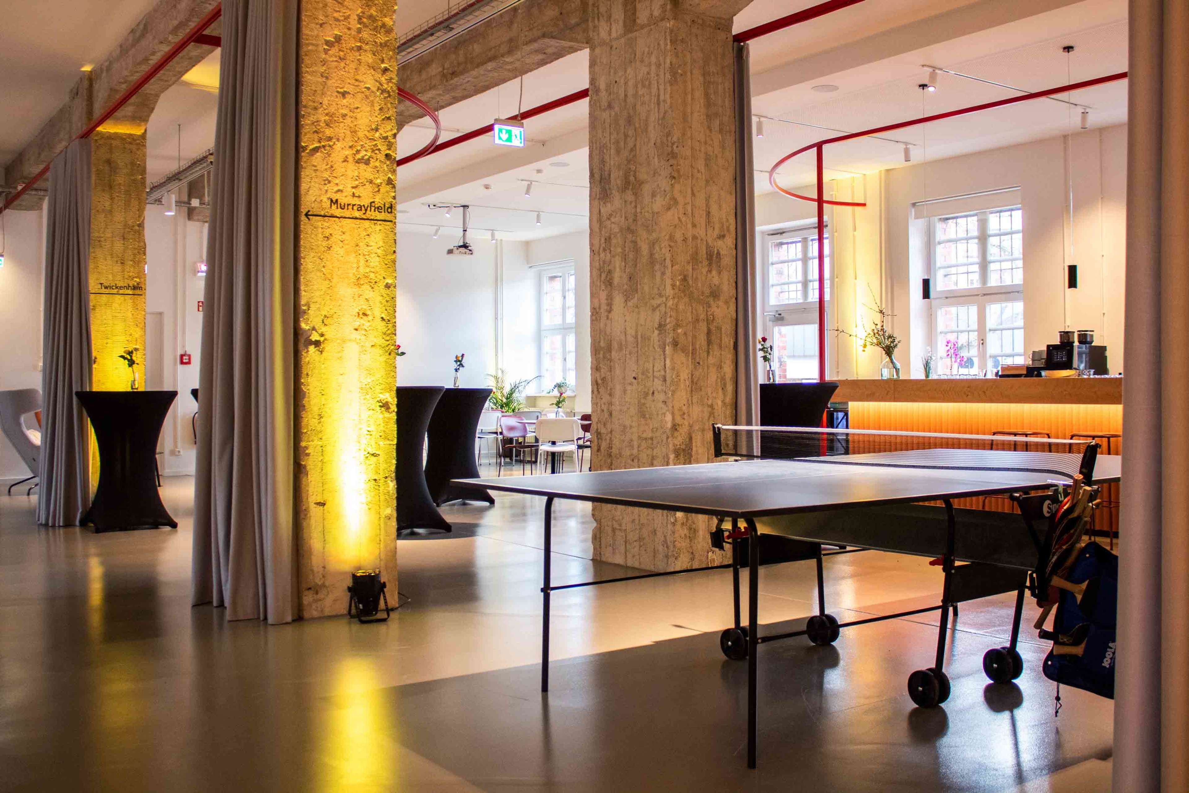 A table tennis setup is positioned in a spacious, modern interior with concrete pillars and a bar area in the background.