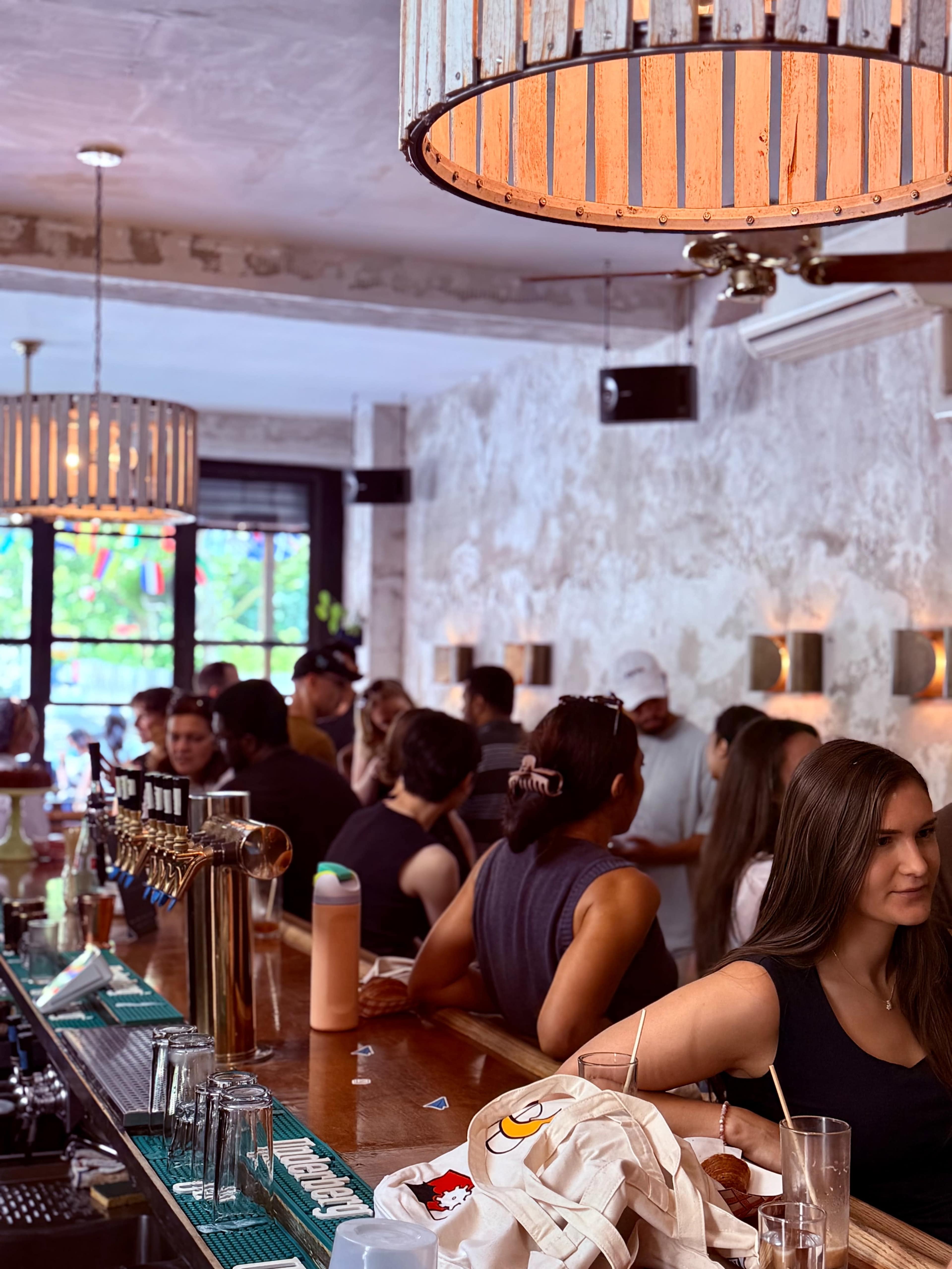 The image shows a busy bar with a long wooden counter, hanging lights, and a group of people socializing in a well-lit, stylish interior.