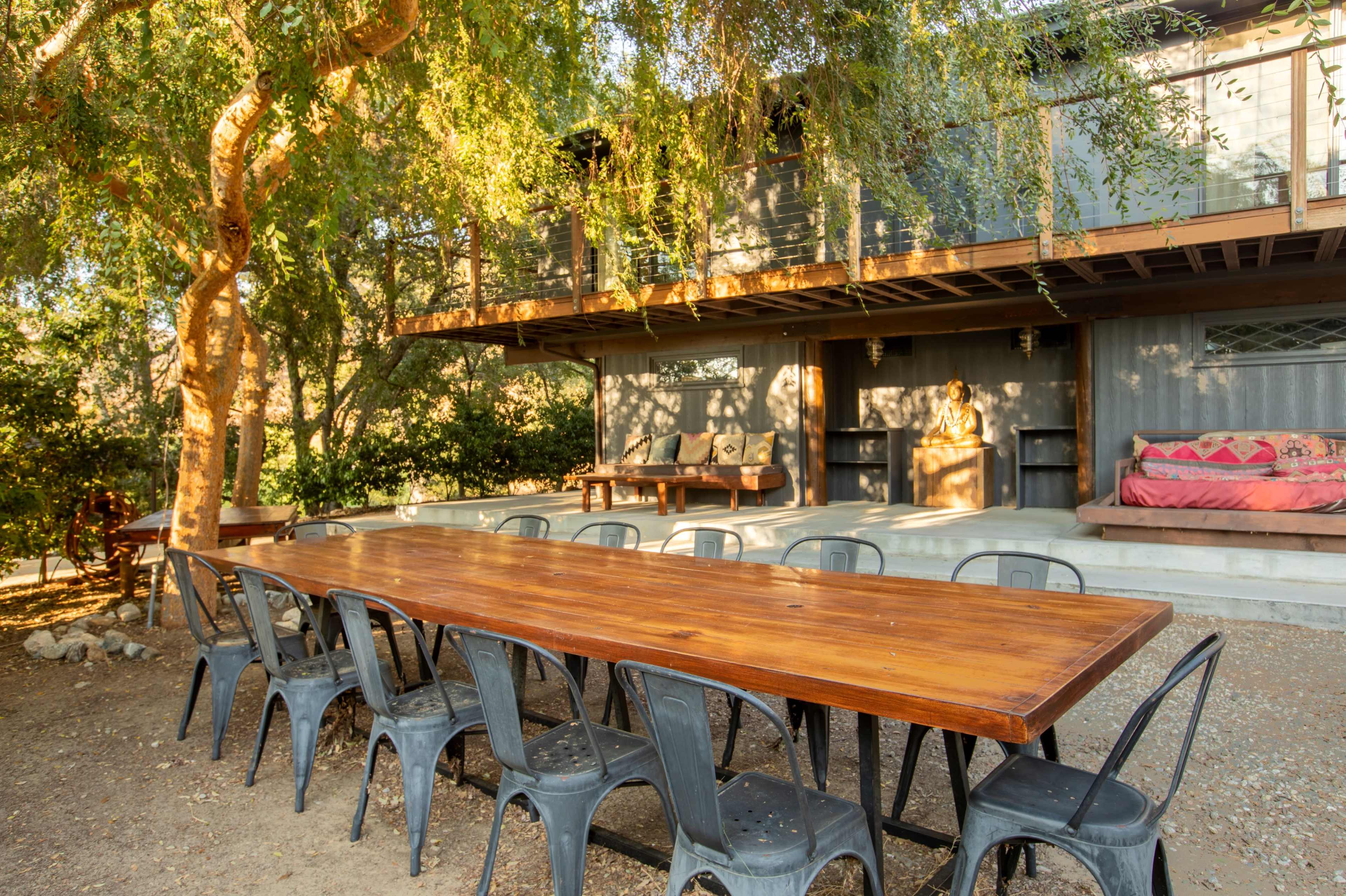 A long wooden dining table with metal chairs is set outside under a tree, in front of a two-story house with a balcony.
