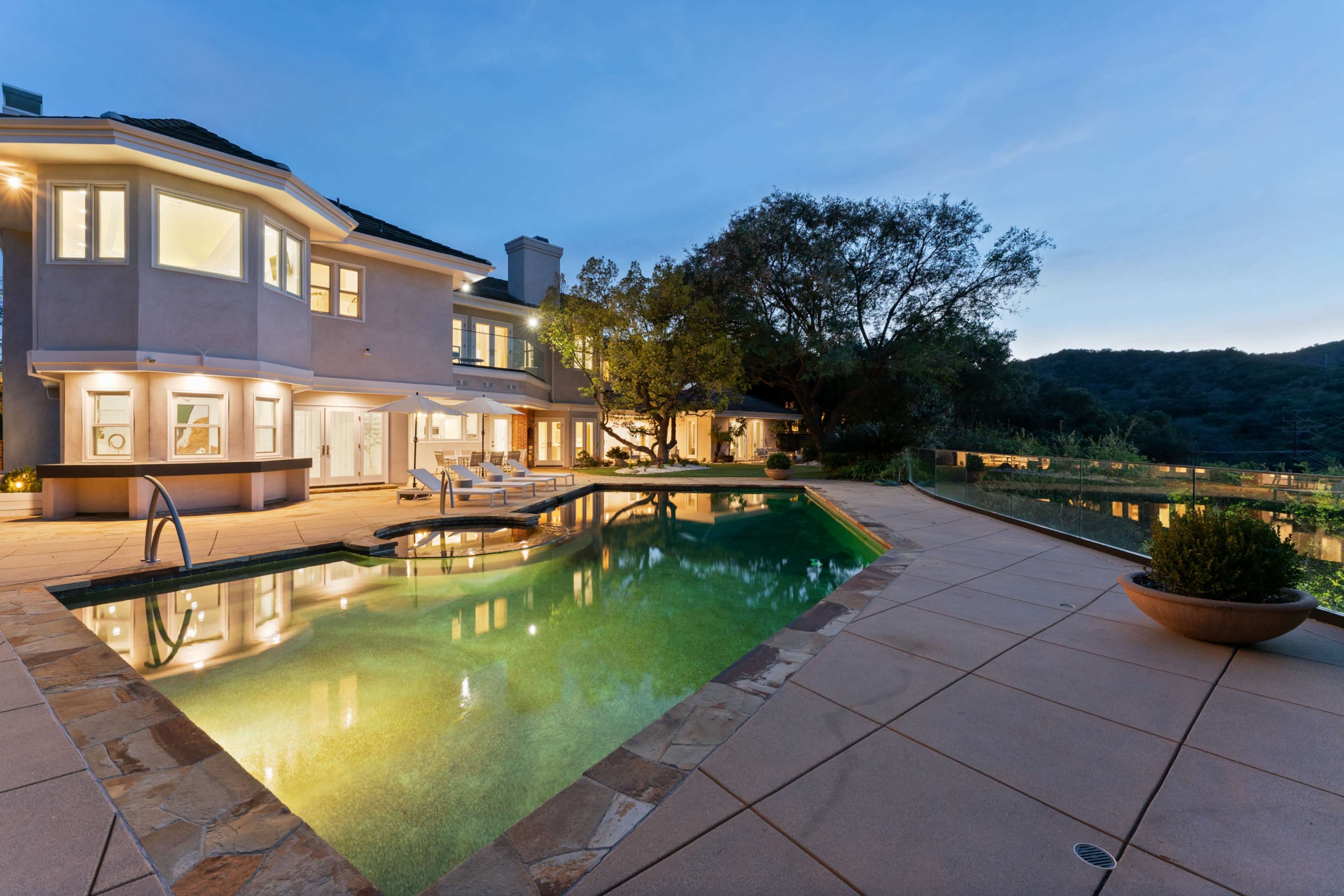 The image shows a spacious modern home with a swimming pool surrounded by a tiled patio, set against a backdrop of rolling hills at dusk.