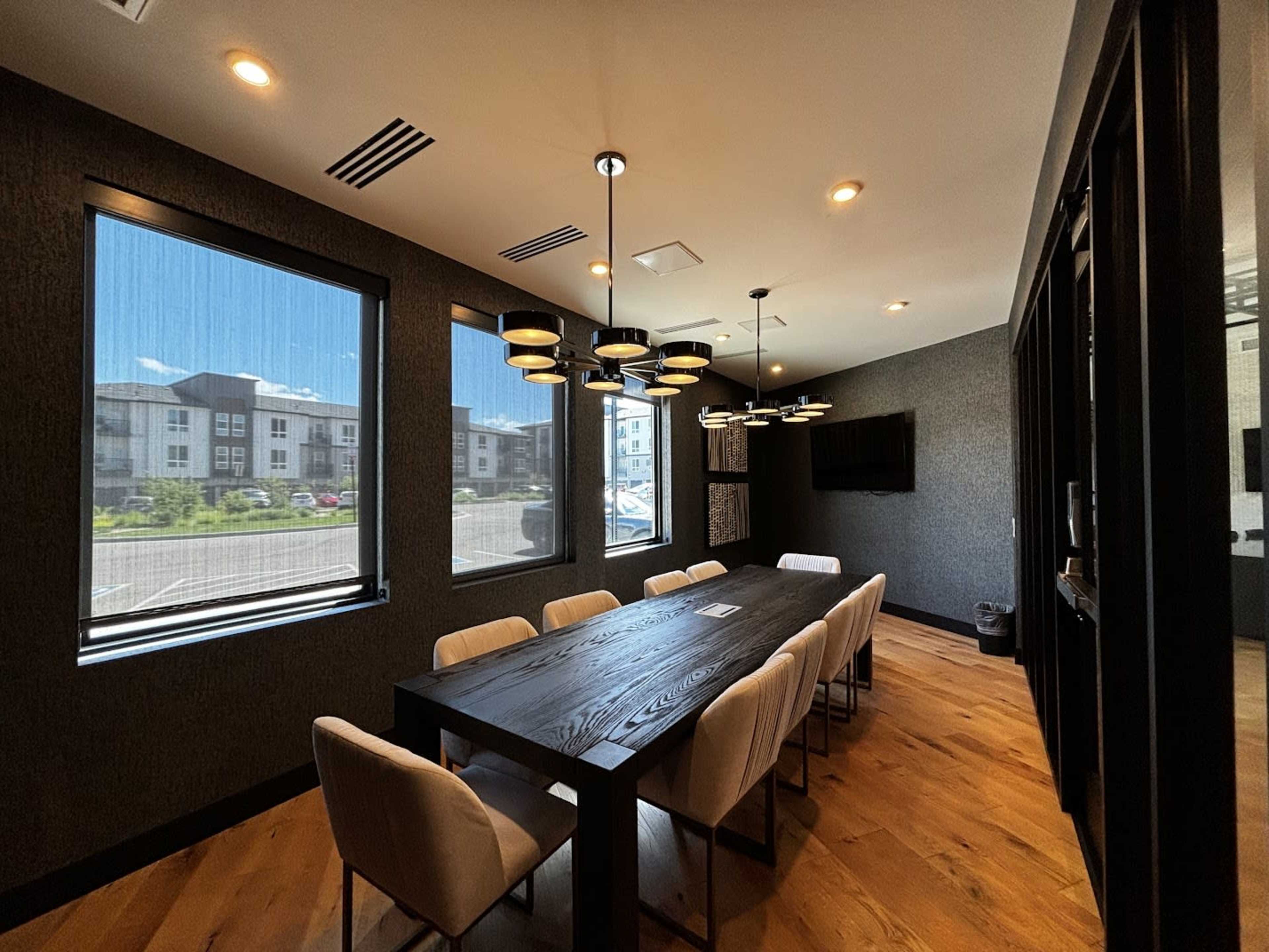 A modern conference room features a large black table surrounded by light-colored chairs, illuminated by pendant lighting and natural light through large windows.