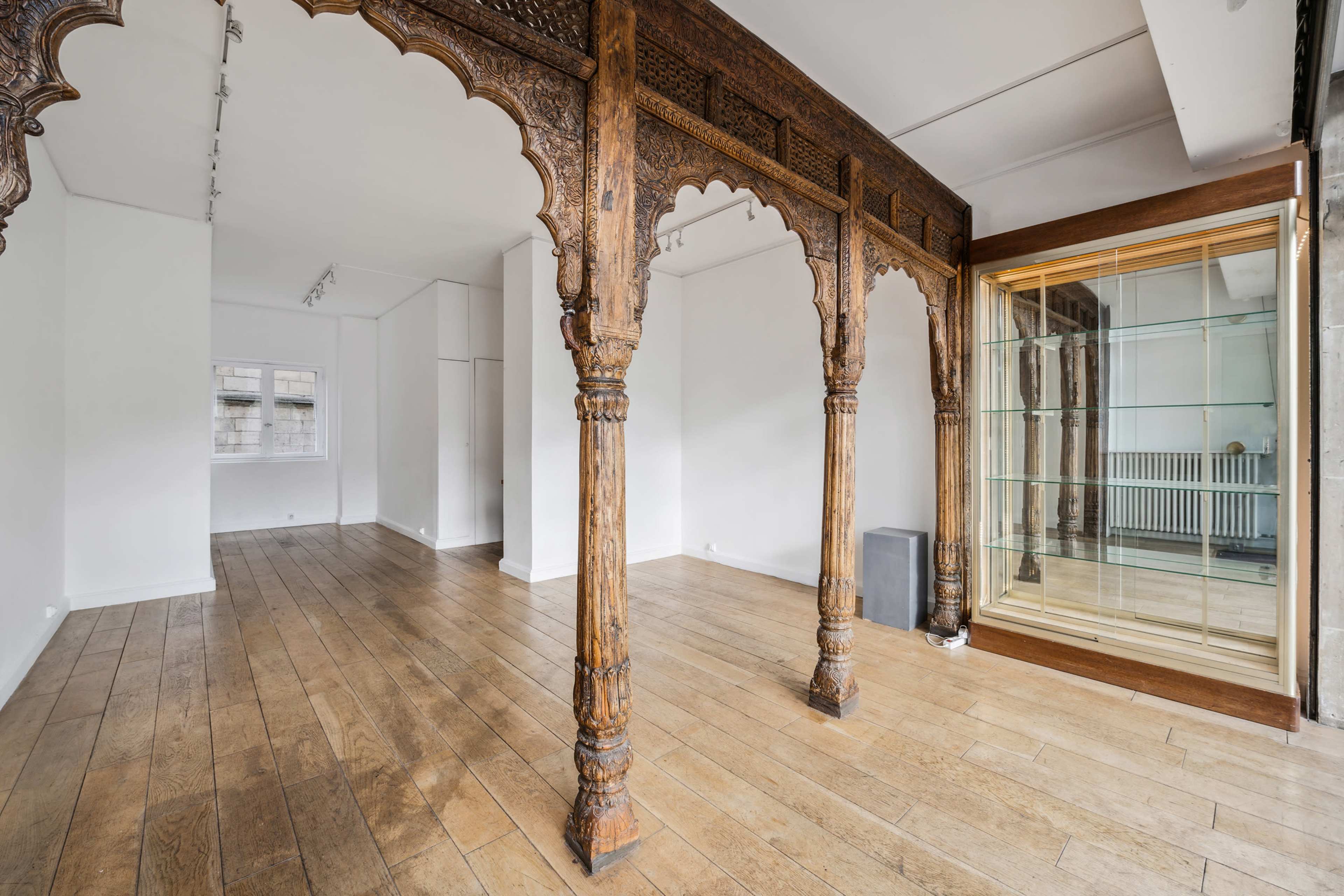 The image shows a spacious room with wooden flooring and intricately carved wooden pillars, alongside a glass display cabinet.