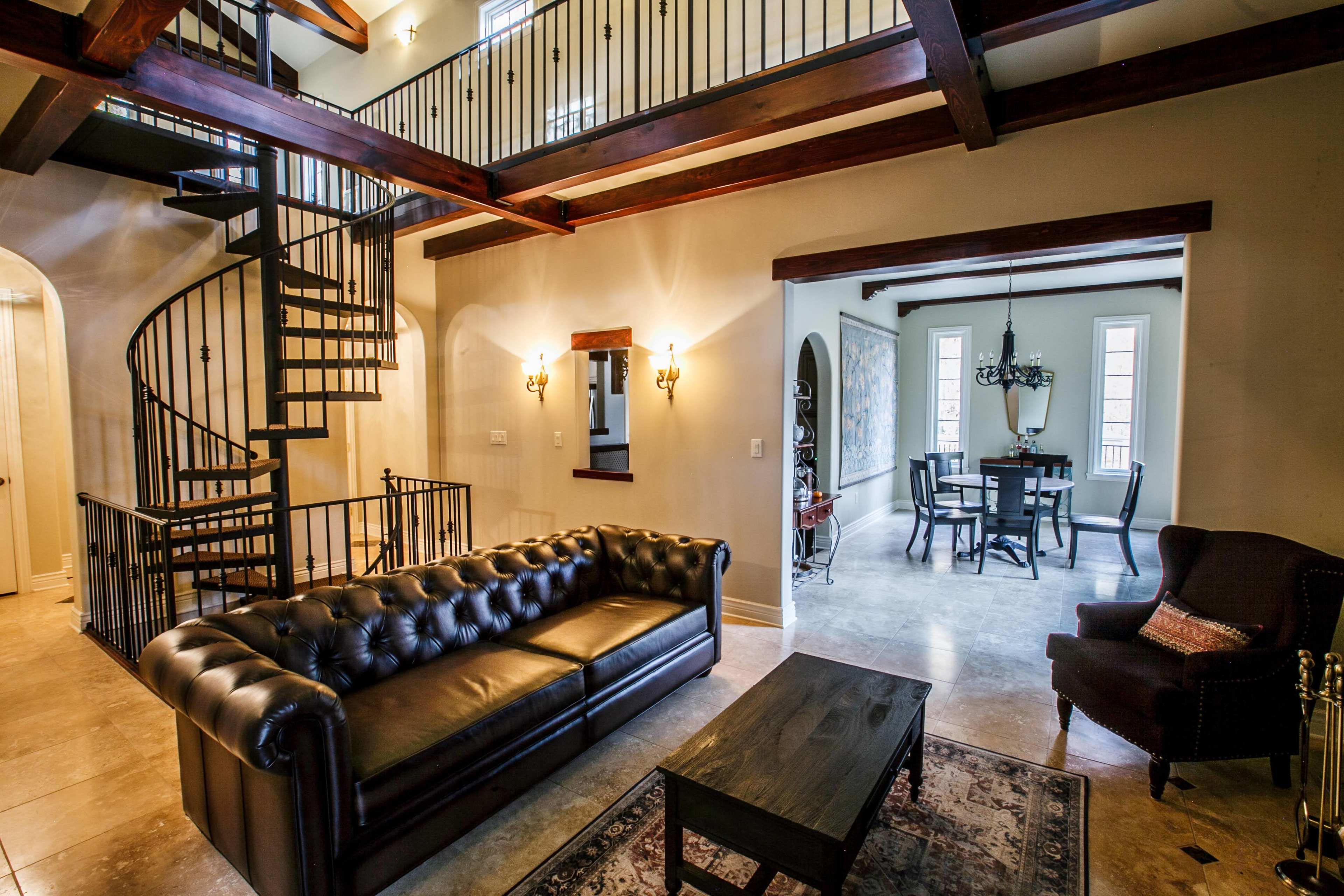 A spacious living area with a black leather sofa, a coffee table, and a spiral staircase leading to a second level, with a dining area visible in the background.