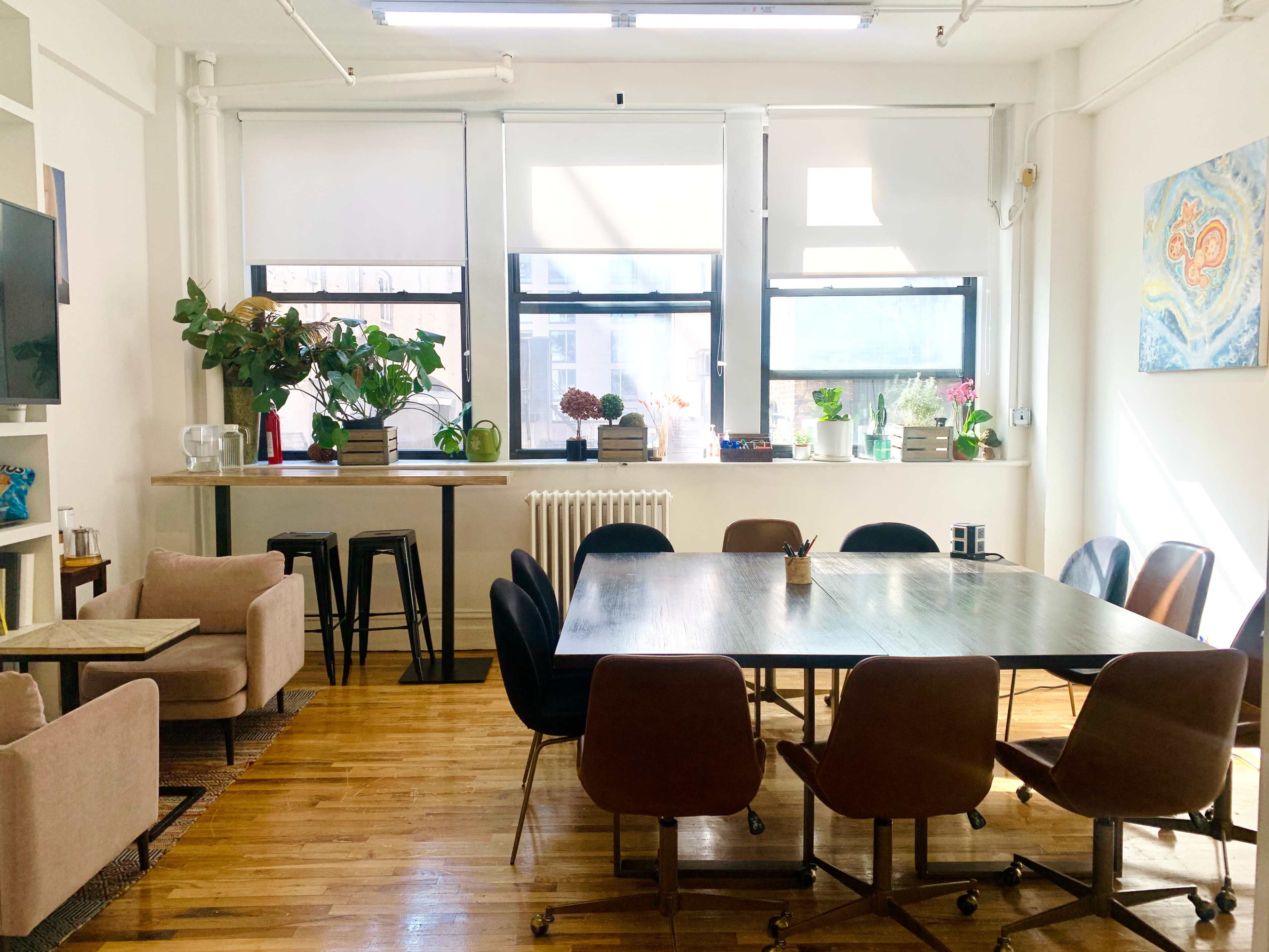 A bright, open room features a large table surrounded by chairs, with a small seating area and plants near the windows.