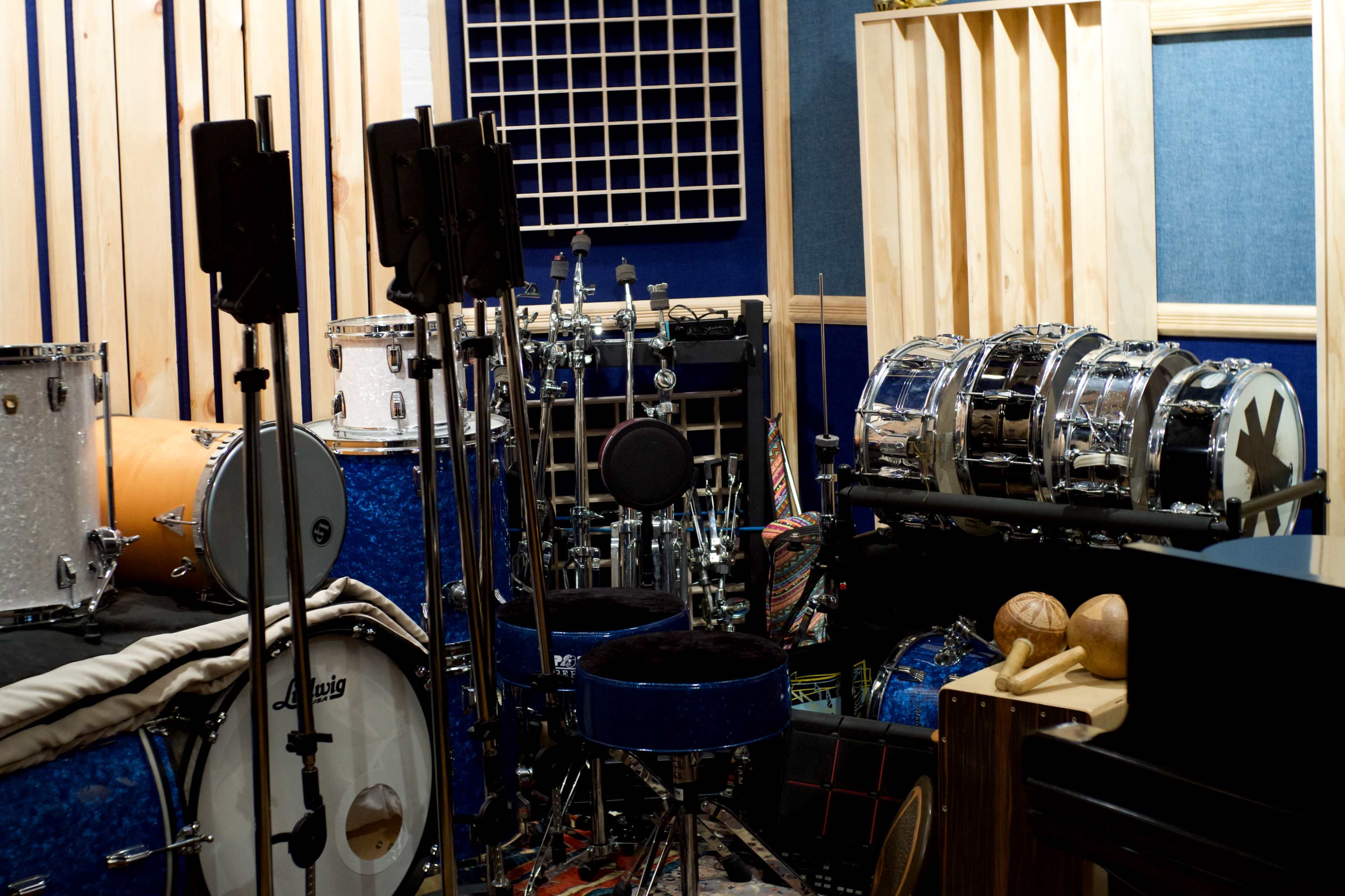 The image shows a recording studio filled with various drums, microphones, and wooden panels for sound treatment.