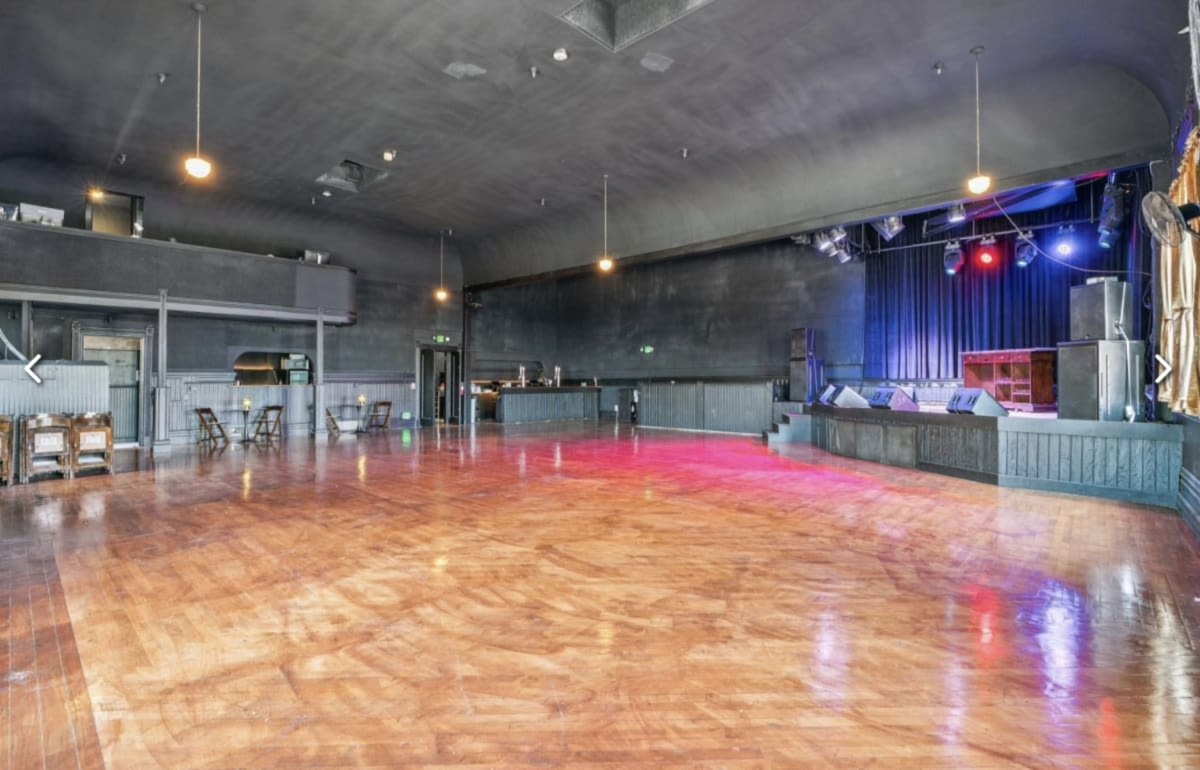 The image shows a spacious performance venue with a polished wooden floor, a stage at the front, and seating arrangements along the sides.