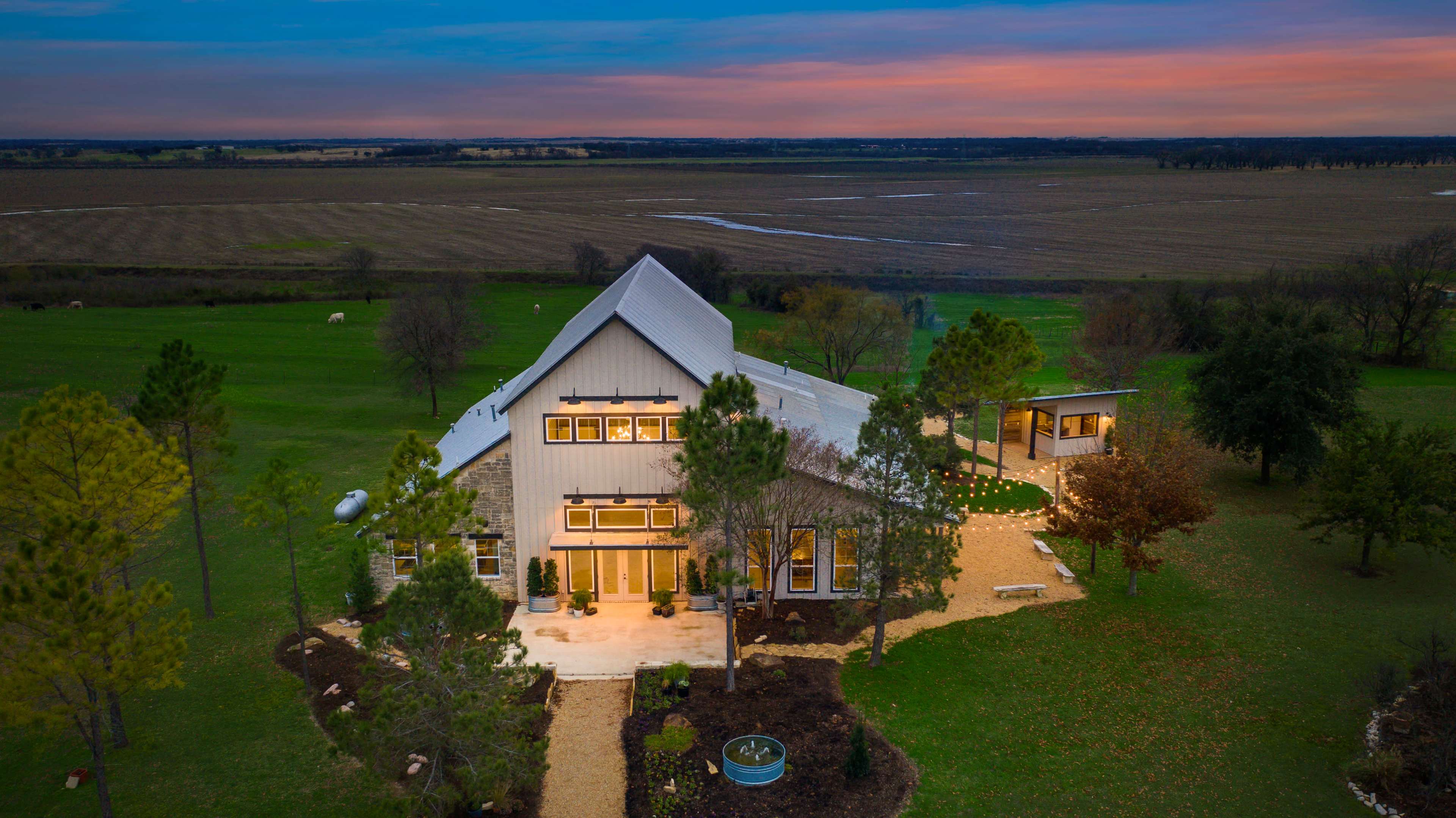 The image shows a modern house with a gabled roof situated in a large green landscape during sunset, with farmland visible in the background.