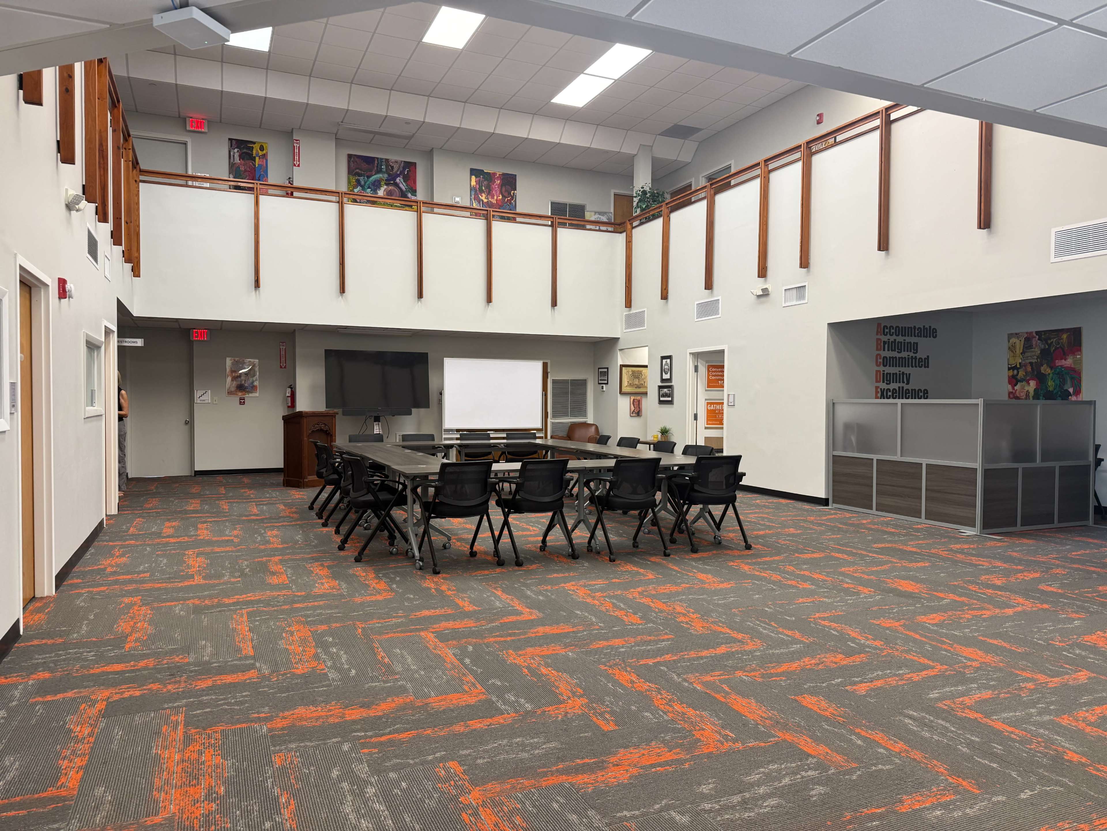 The image shows a spacious classroom or meeting area with a large table set up for discussion, surrounded by black chairs, and decorated with wall art and various educational materials.