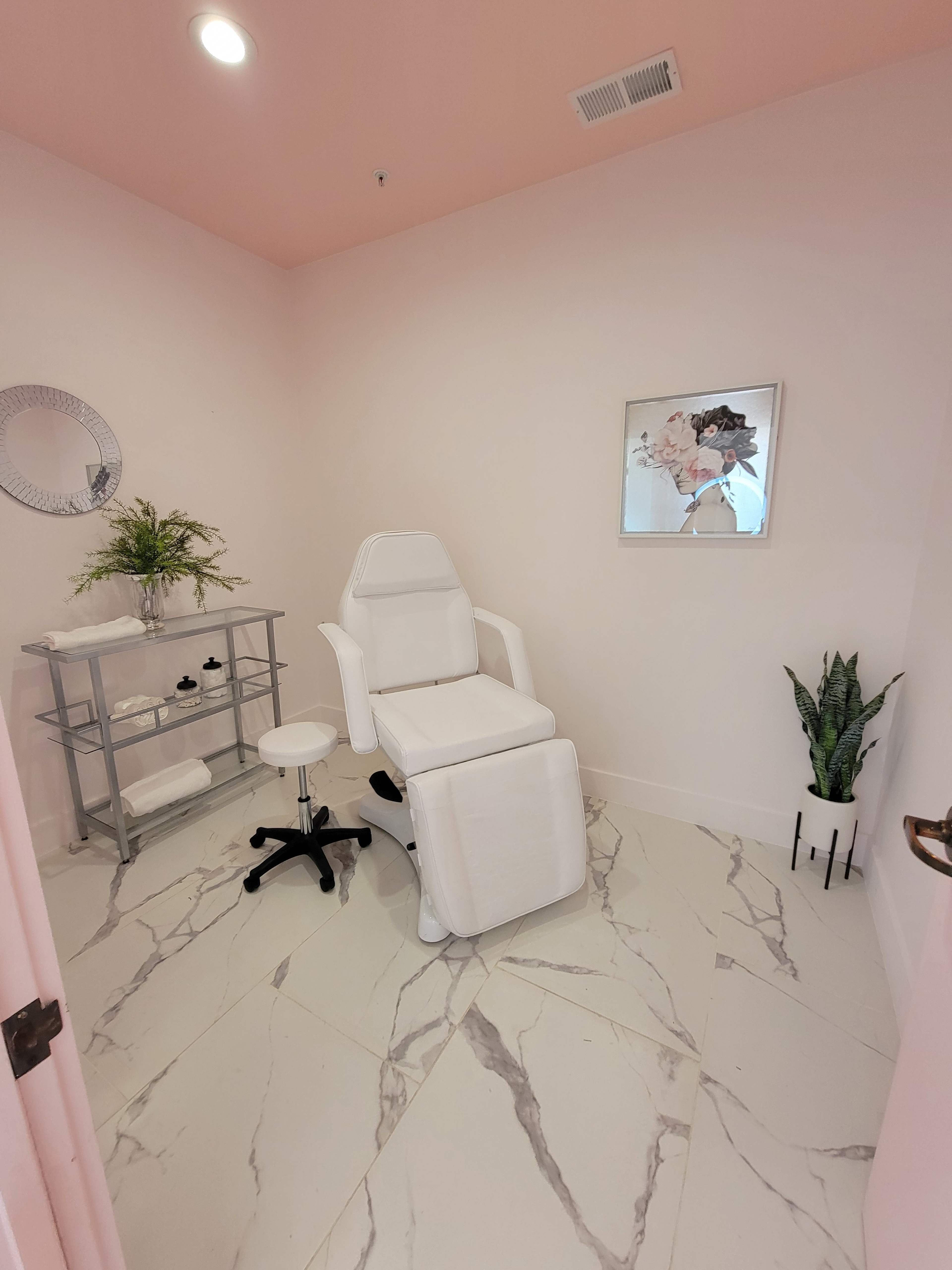 A bright, minimalist treatment room featuring a white reclining chair, a small rolling stool, a metal cart with items, and a potted plant in the corner.