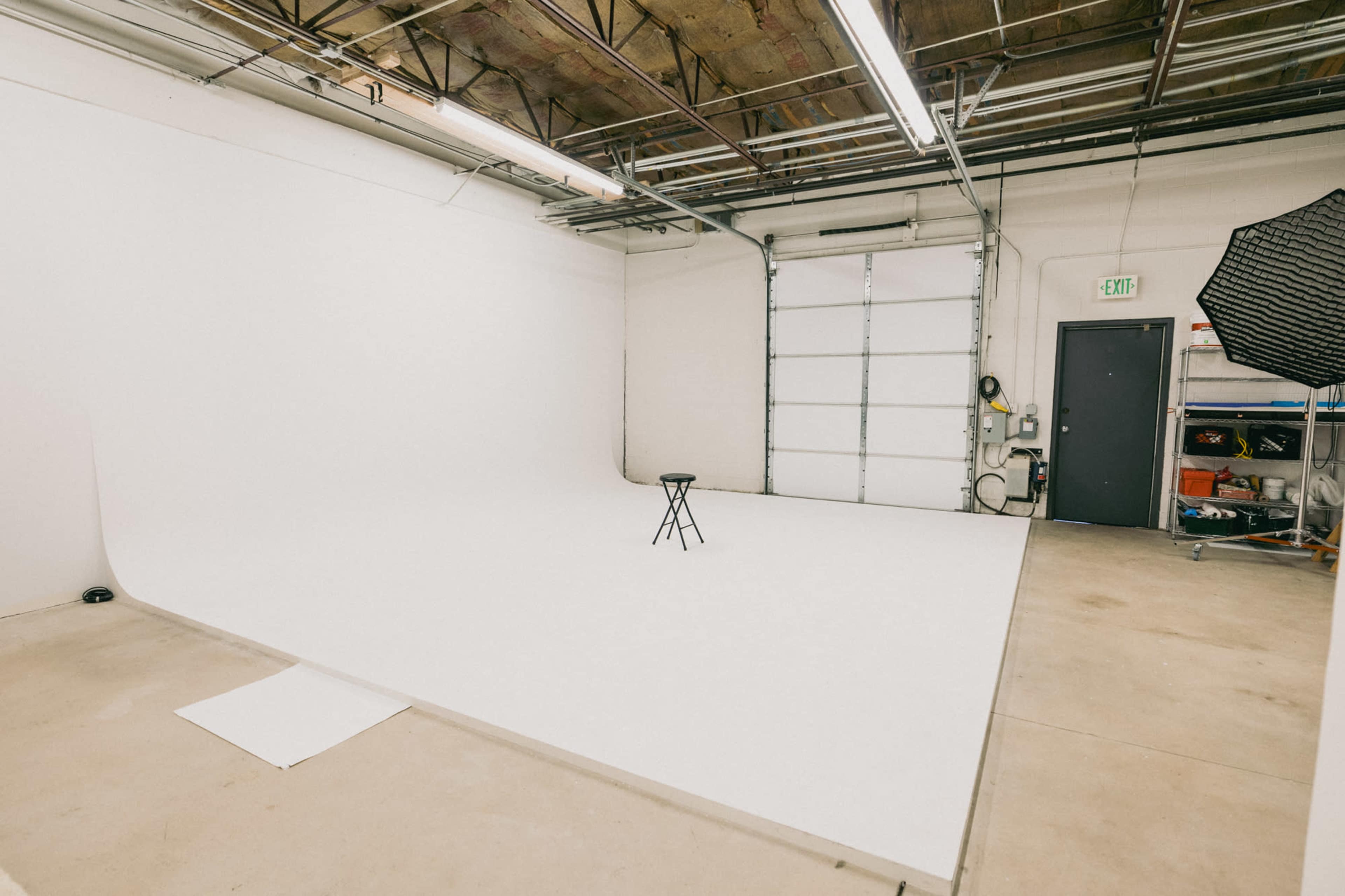 The image shows a spacious photo studio with a seamless white backdrop, a stool positioned in the center, and various equipment stored along the walls.