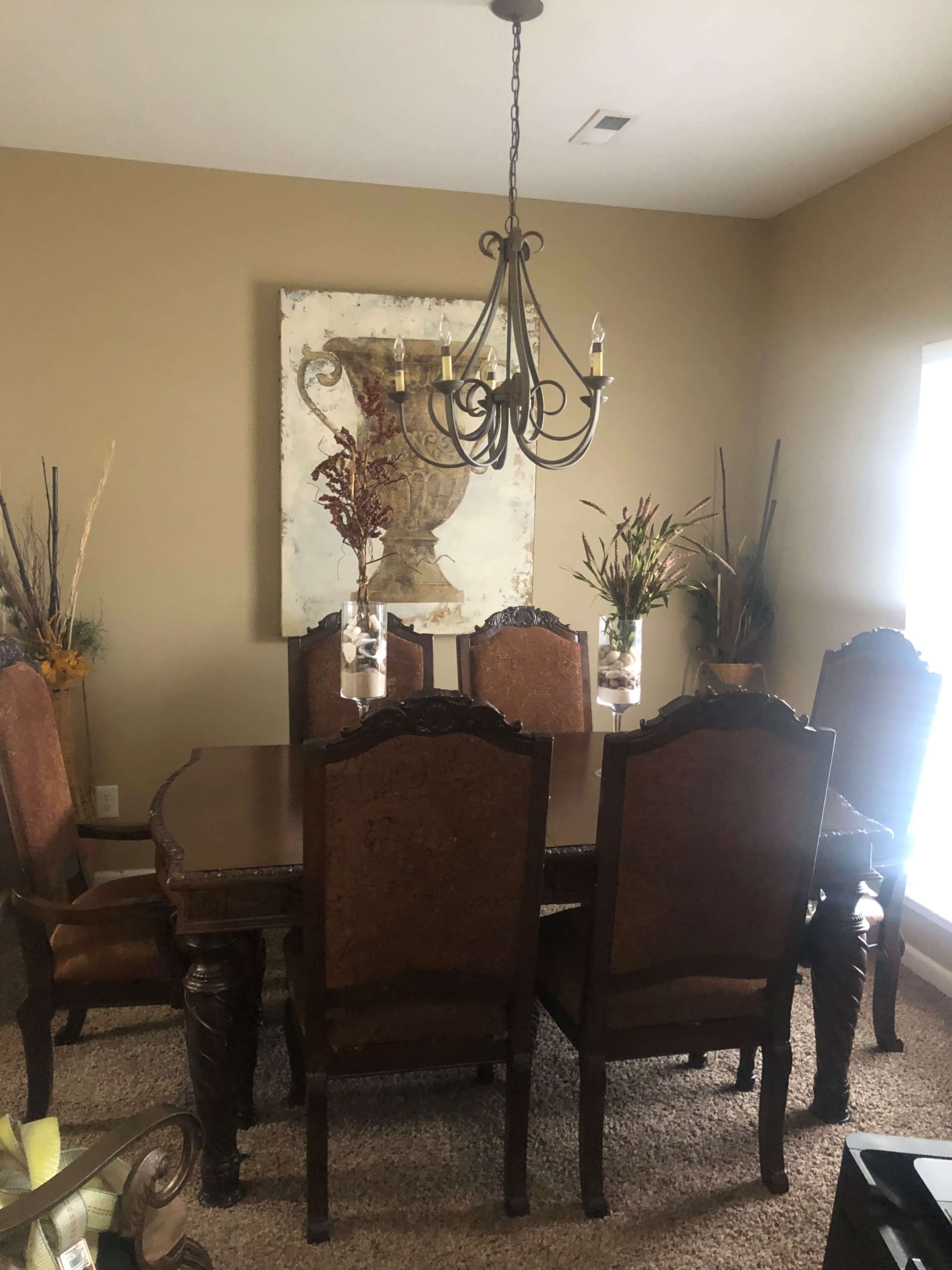 A dining room features a large wooden table surrounded by upholstered chairs, with a chandelier above and a decorative painting on the wall.