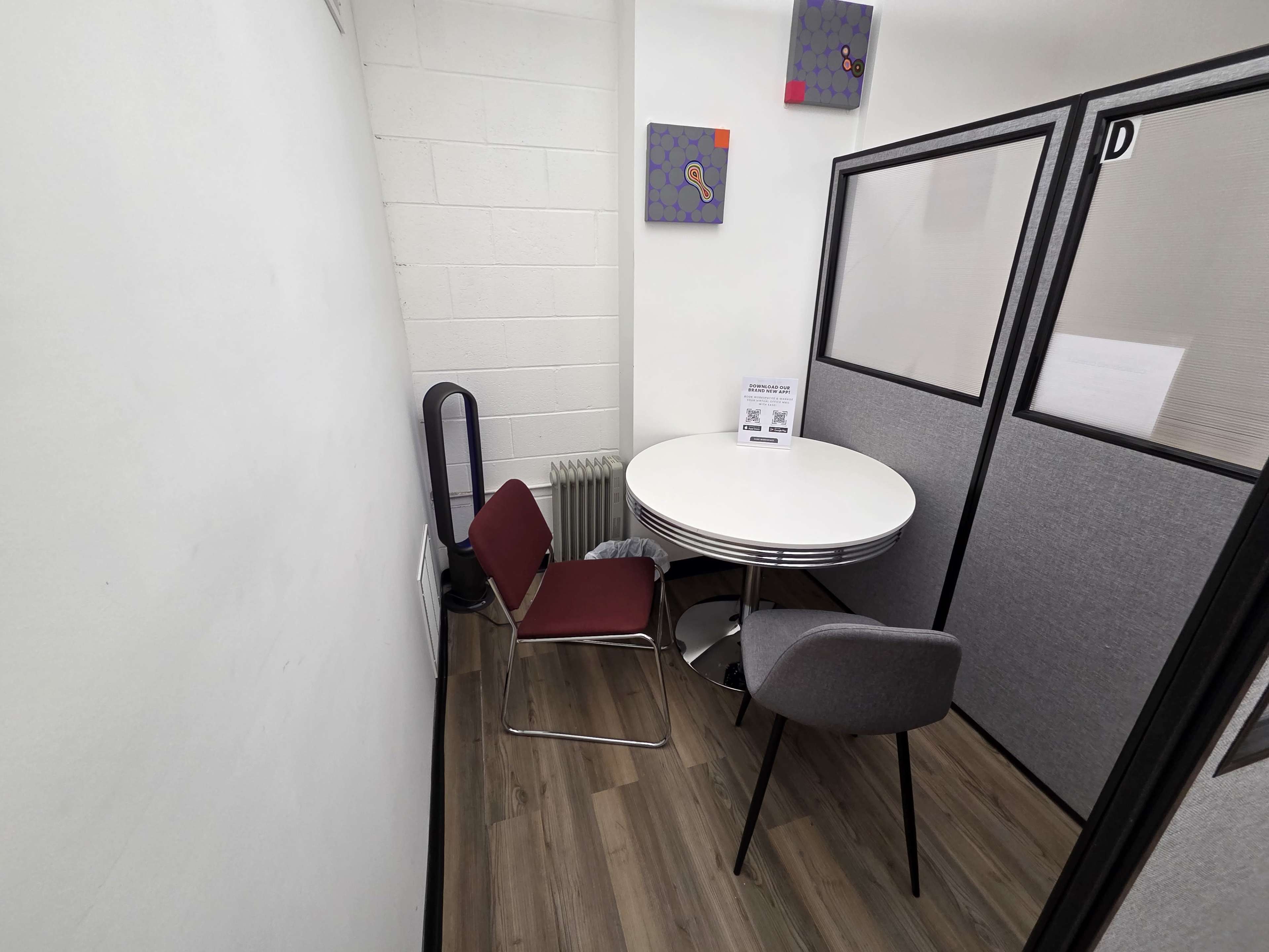 The image shows a small corner office space with a round white table, two chairs, and a partitioned area featuring gray panels and artwork on the walls.