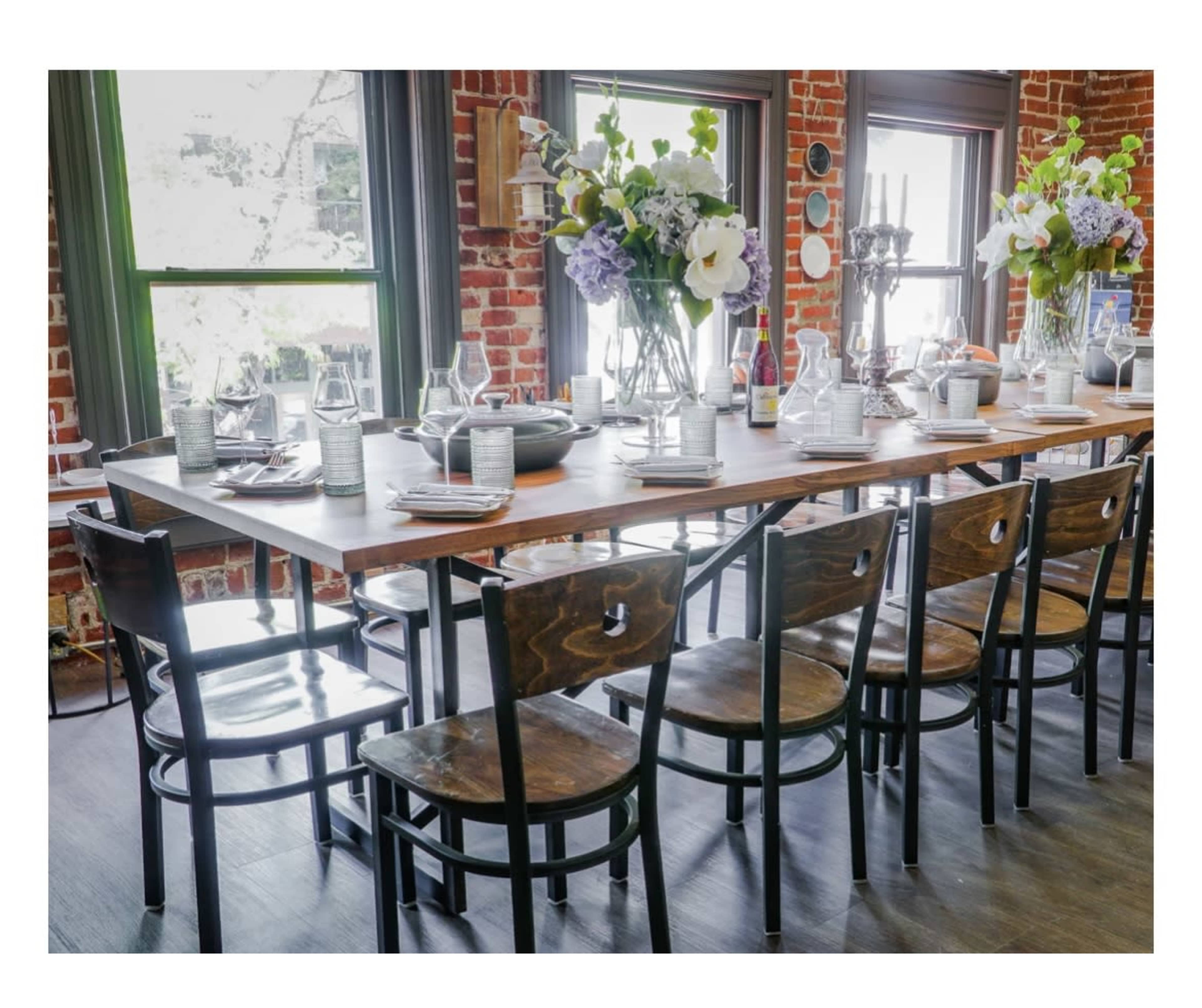 A wooden dining table is set with dishes and glassware, surrounded by black wooden chairs, in a room featuring exposed brick walls and large windows.