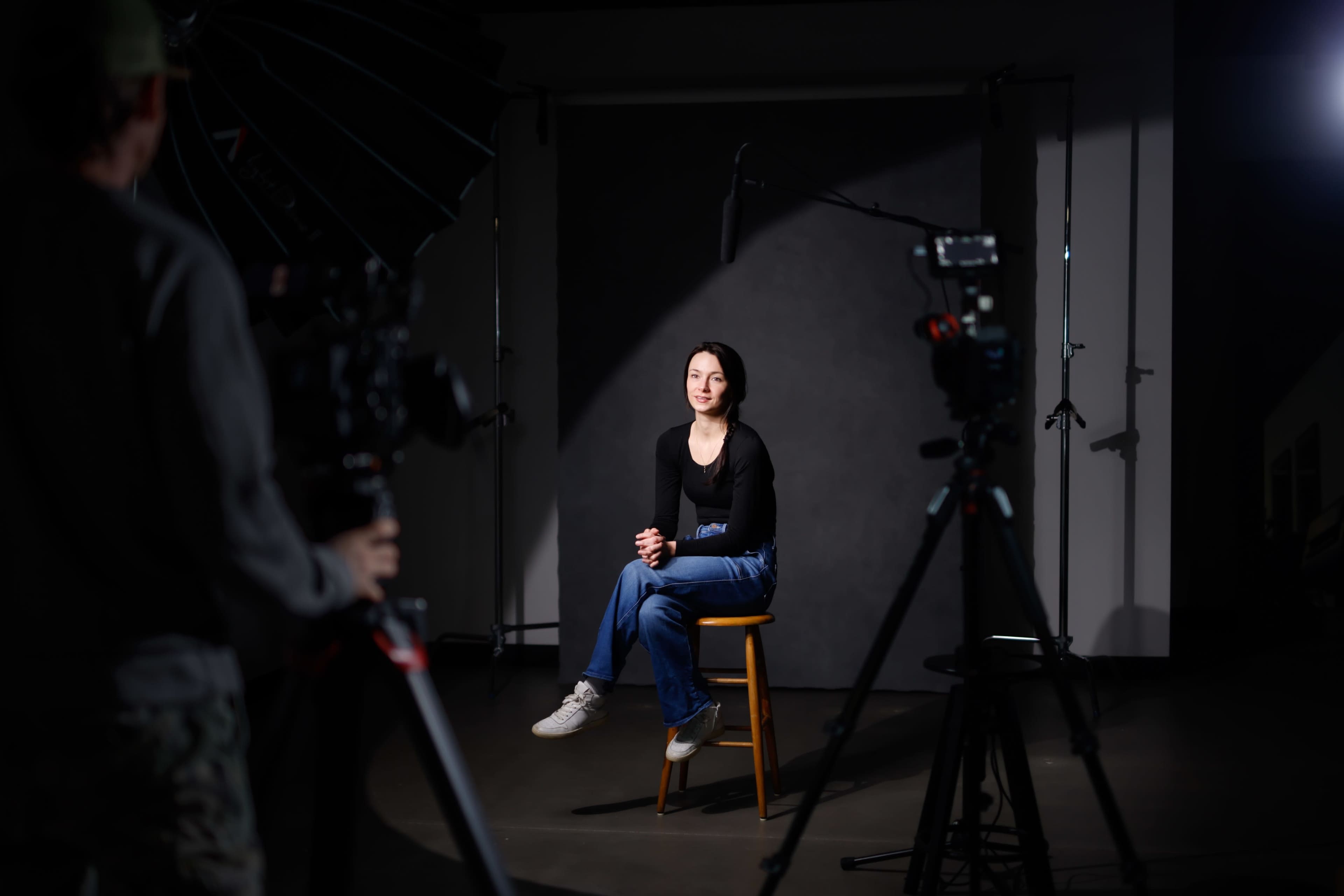 A woman sits on a stool in front of a gray backdrop, illuminated by lights while cameras are set up around her.