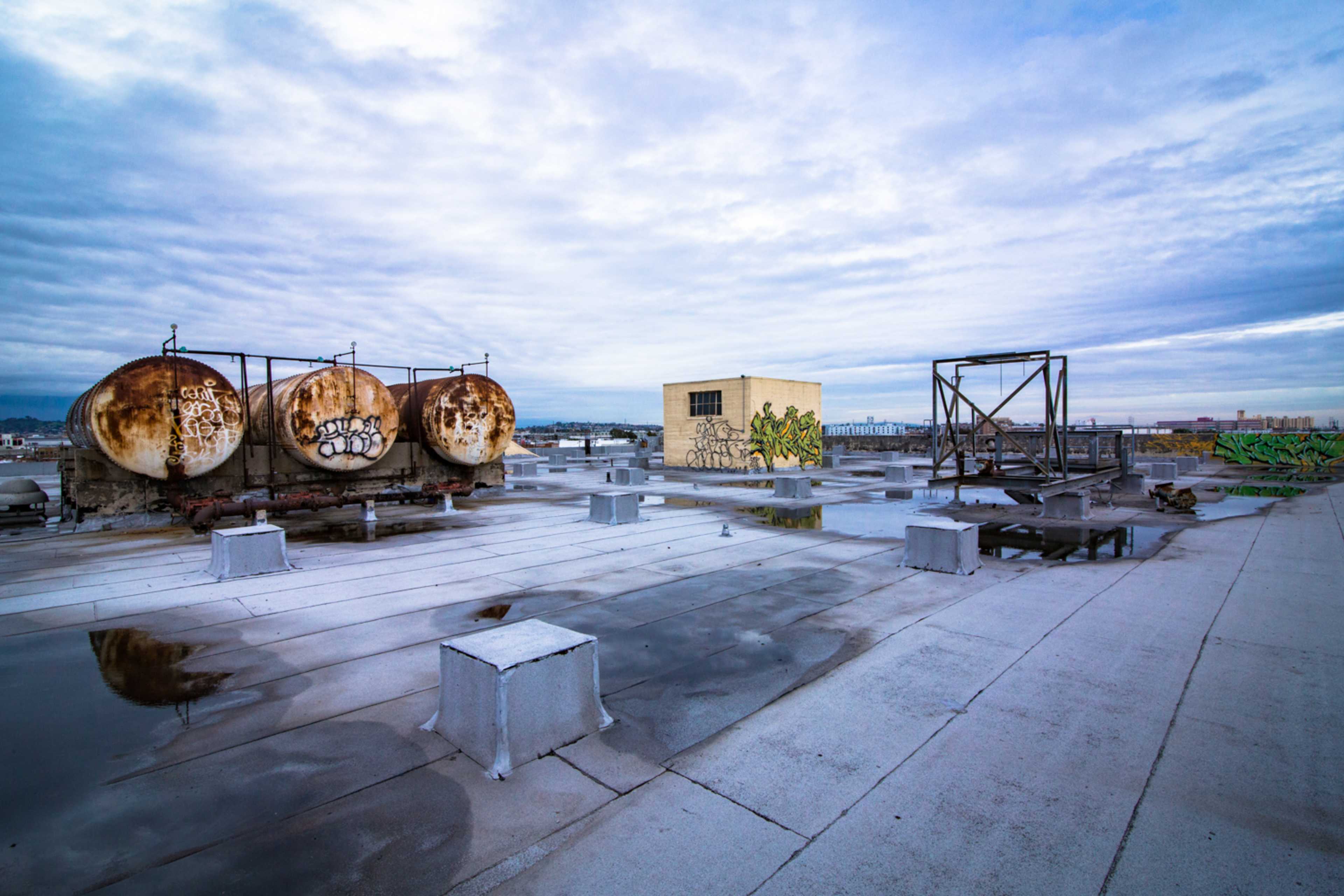 A rooftop with large, weathered storage tanks and a small building, surrounded by puddles and graffiti-covered structures under a cloudy sky.