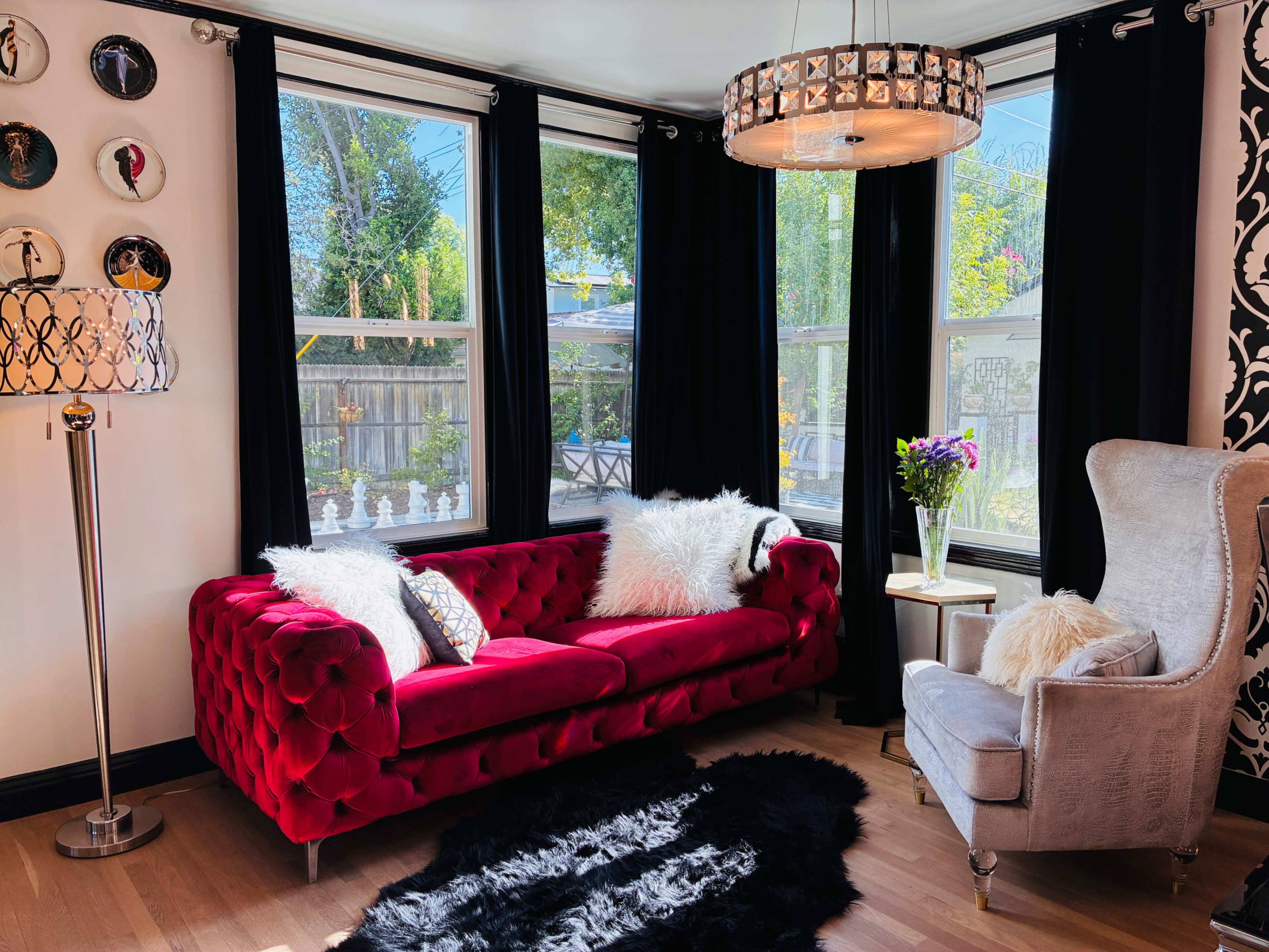 The image shows a cozy living room with a red tufted couch, a gray armchair, a decorative floor lamp, and large windows allowing natural light to enter the space.