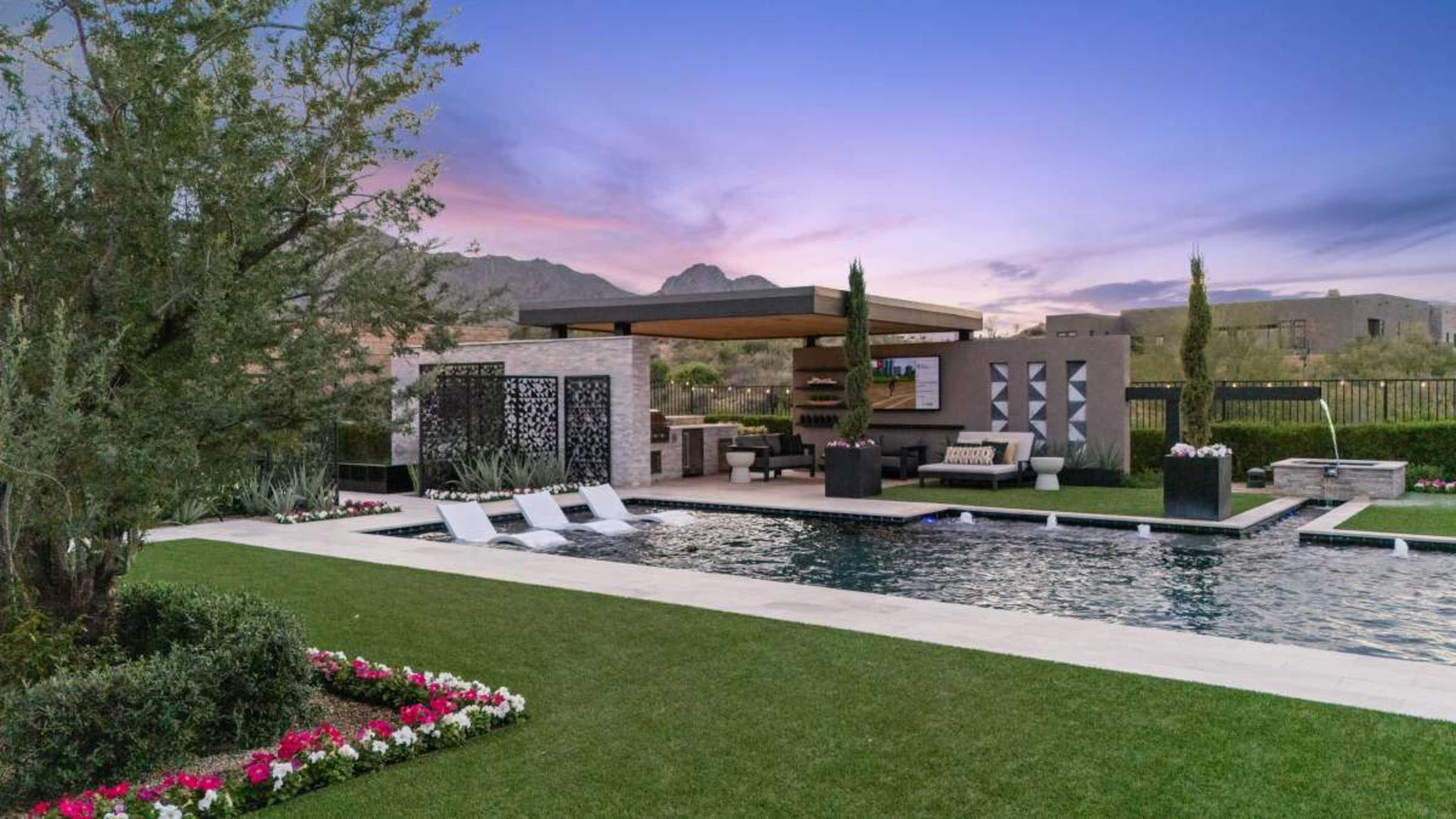 The image shows a modern outdoor pool area with lounge chairs, decorative landscaping, and a covered seating space featuring a television and stone accents.