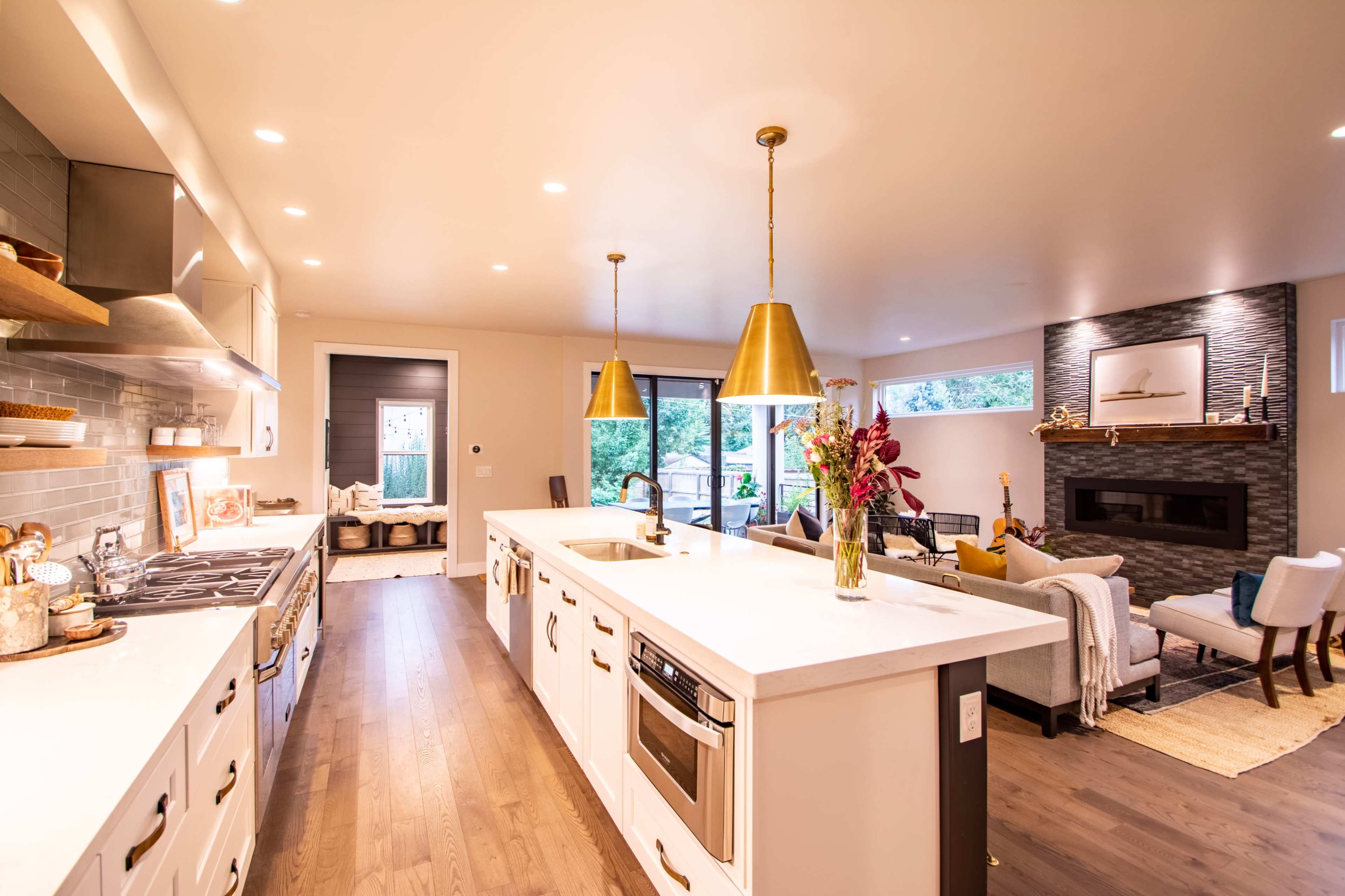 A modern kitchen features white cabinetry, a large island with bar seating, and gold pendant lights, adjoining a living area with a fireplace and large windows.