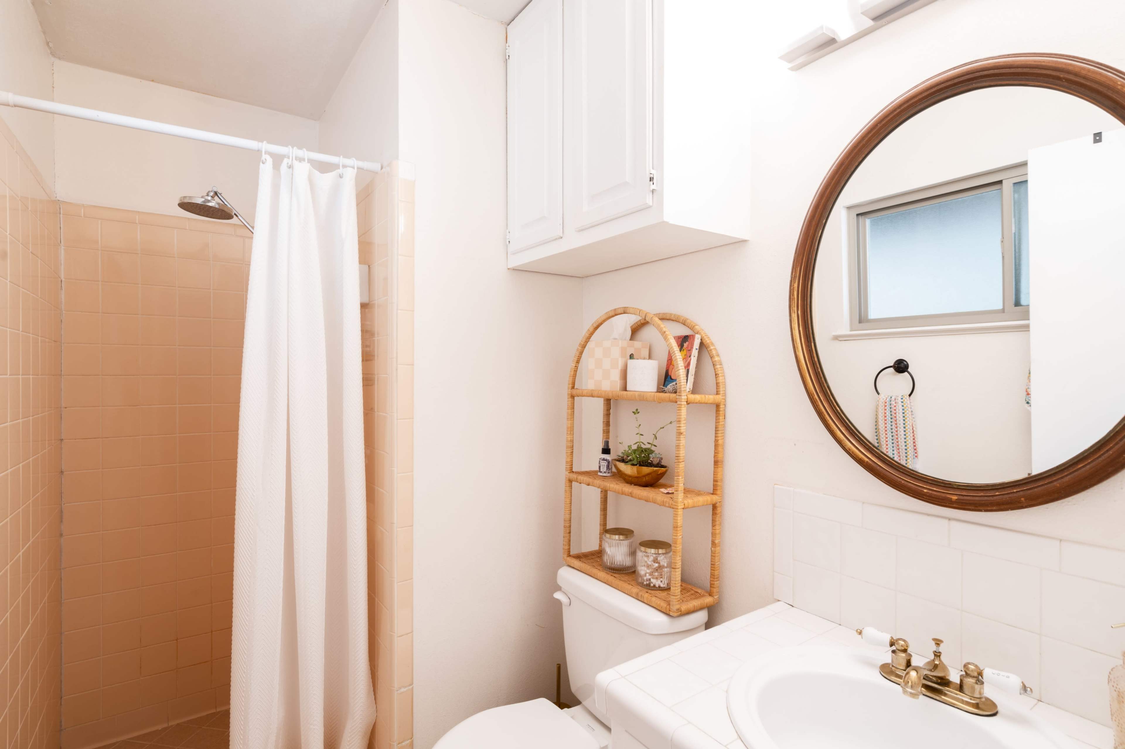 A small bathroom features a shower with a curtain, a round mirror above a sink, and a rattan shelf with plants and decor items.