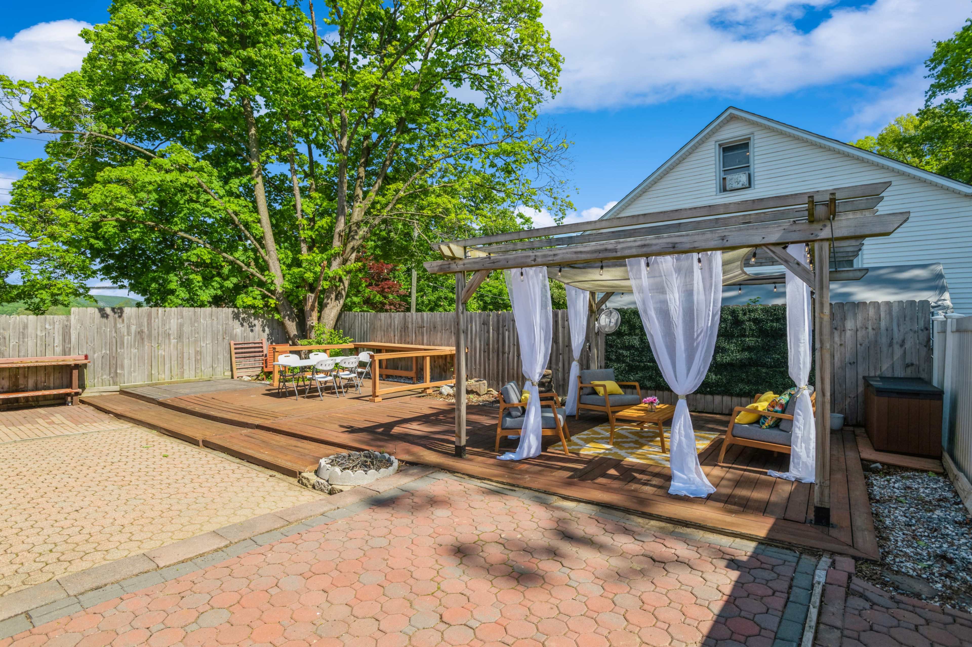 A wooden deck with a pergola and seating area is situated in a backyard surrounded by a wooden fence and large trees.