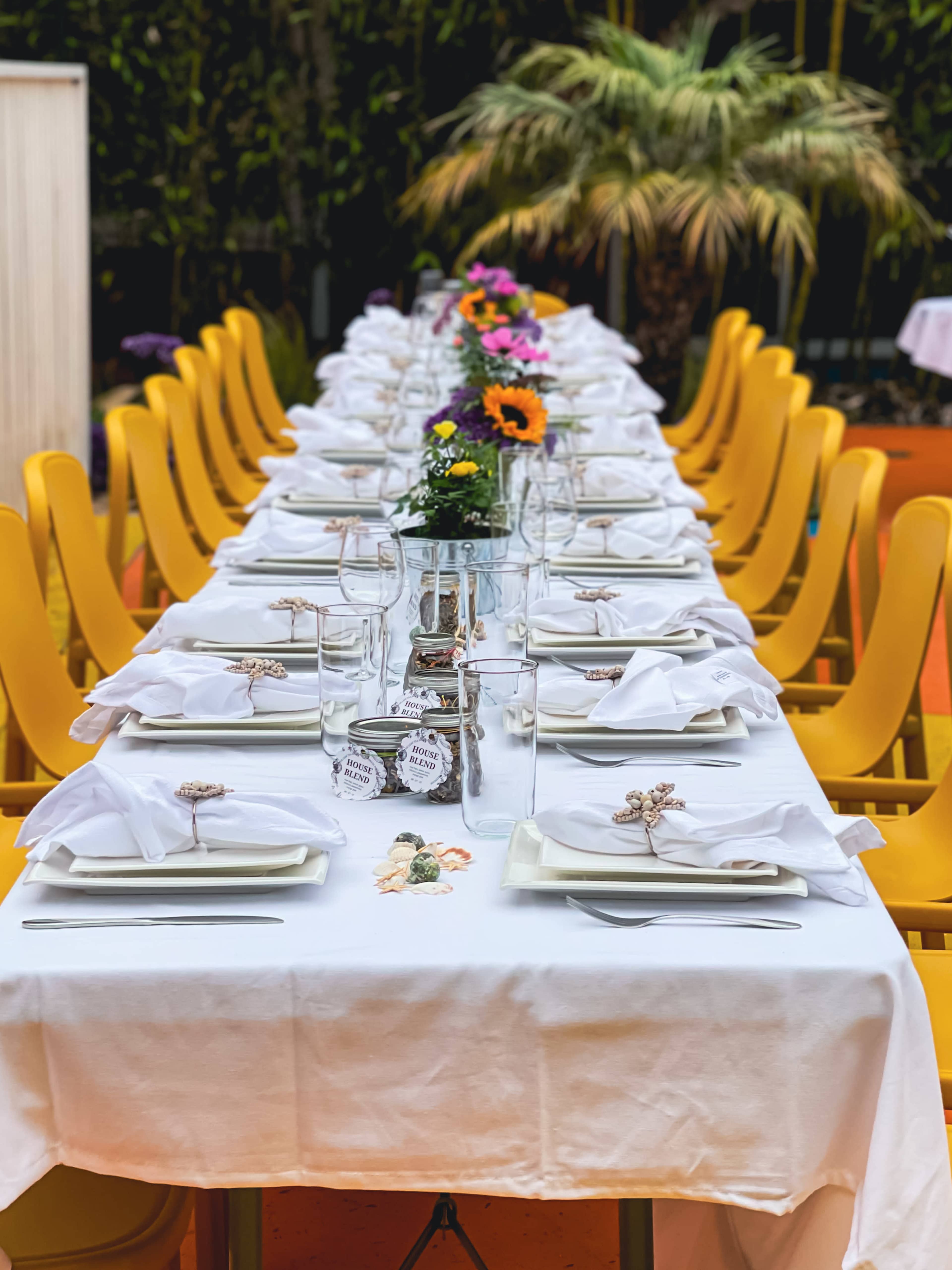 A long dining table is set with white tablecloths, neatly arranged plates, glasses, and small flower centerpieces, surrounded by vibrant yellow chairs.