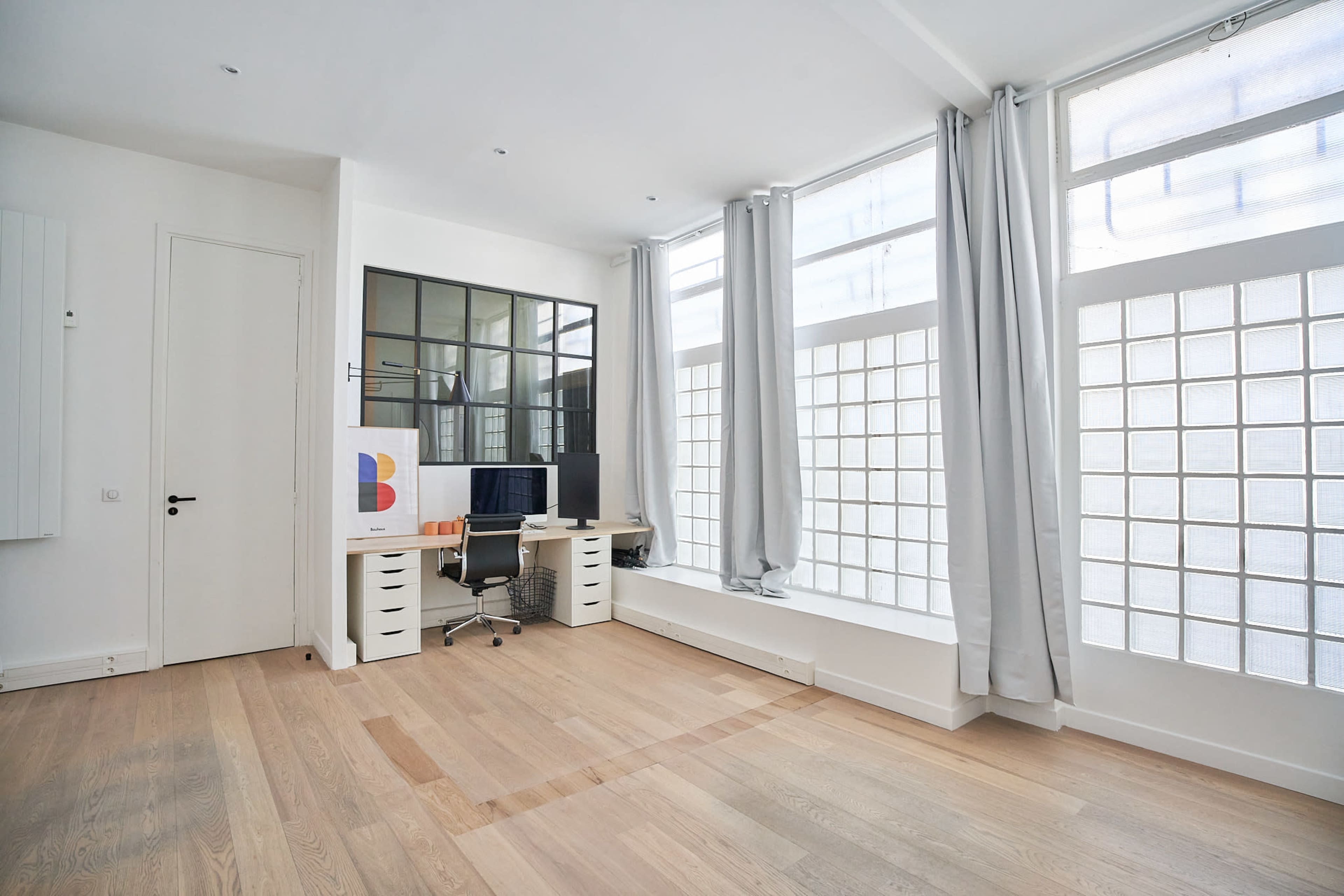 A modern office space with a desk and computer, large glass block windows, and light-colored wooden flooring.