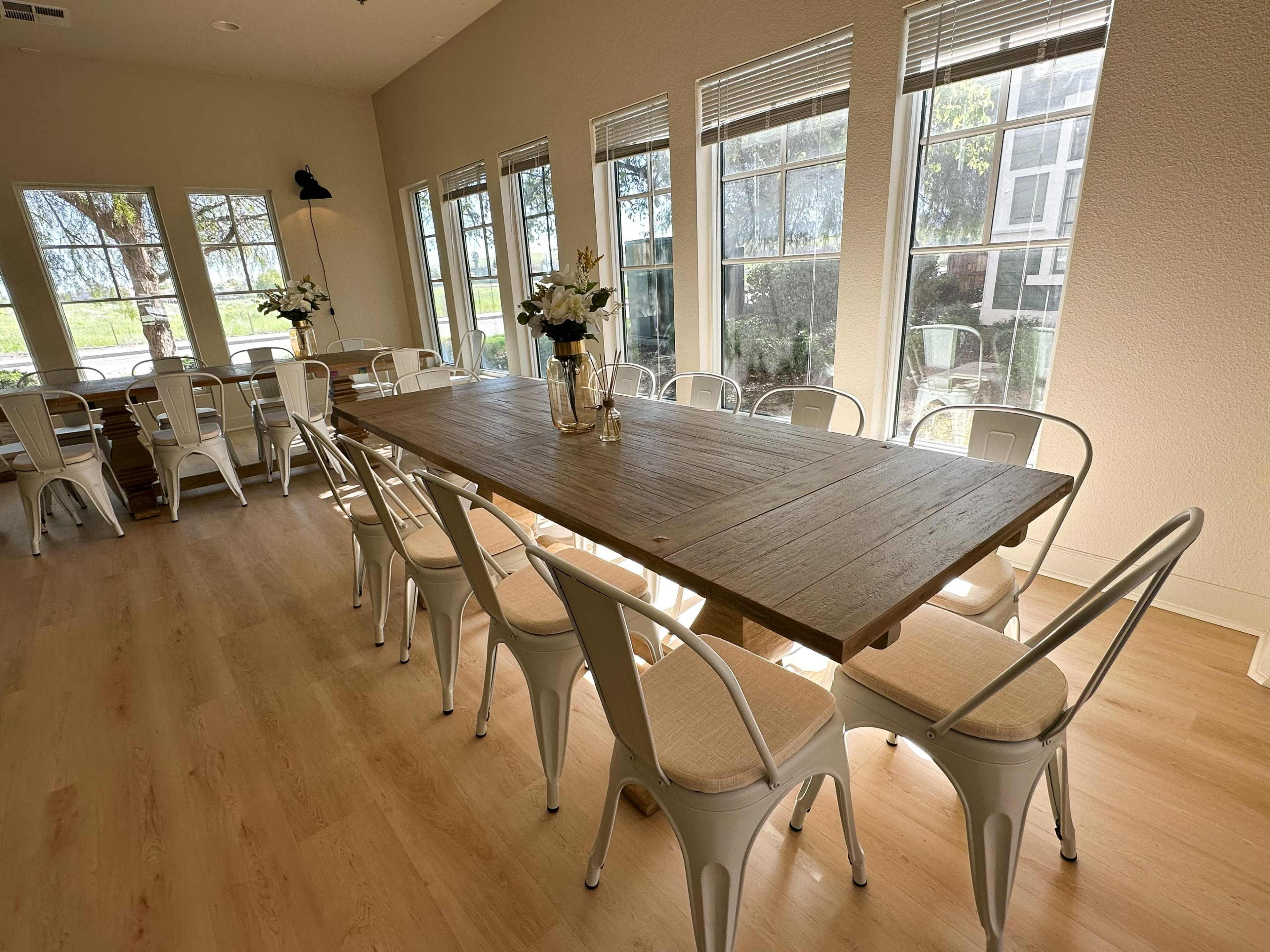 A long wooden table surrounded by white chairs is set in a brightly lit room with large windows.
