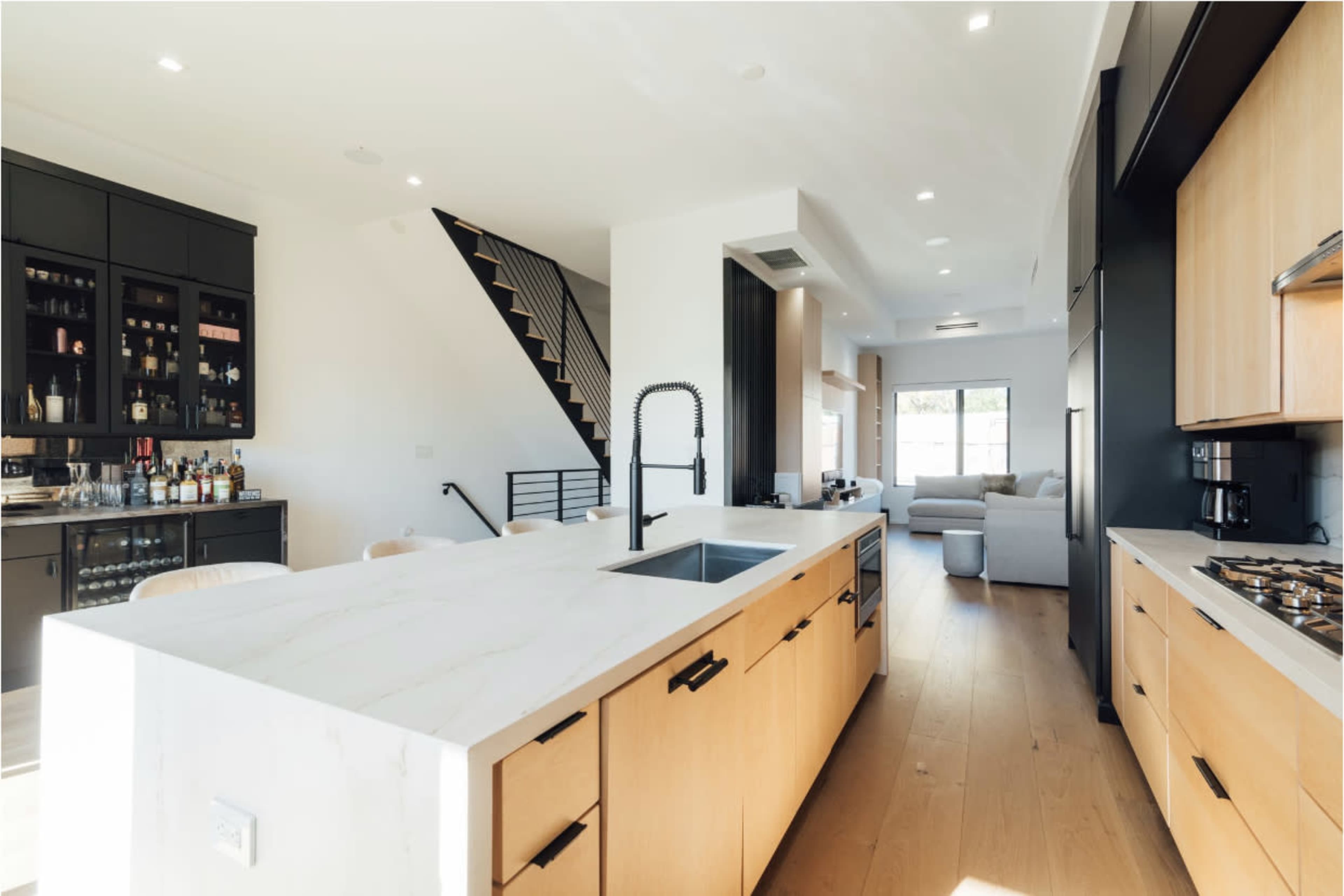 A modern kitchen with a central island, wooden cabinets, and a staircase leading to an upper level, while a living area is visible in the background.