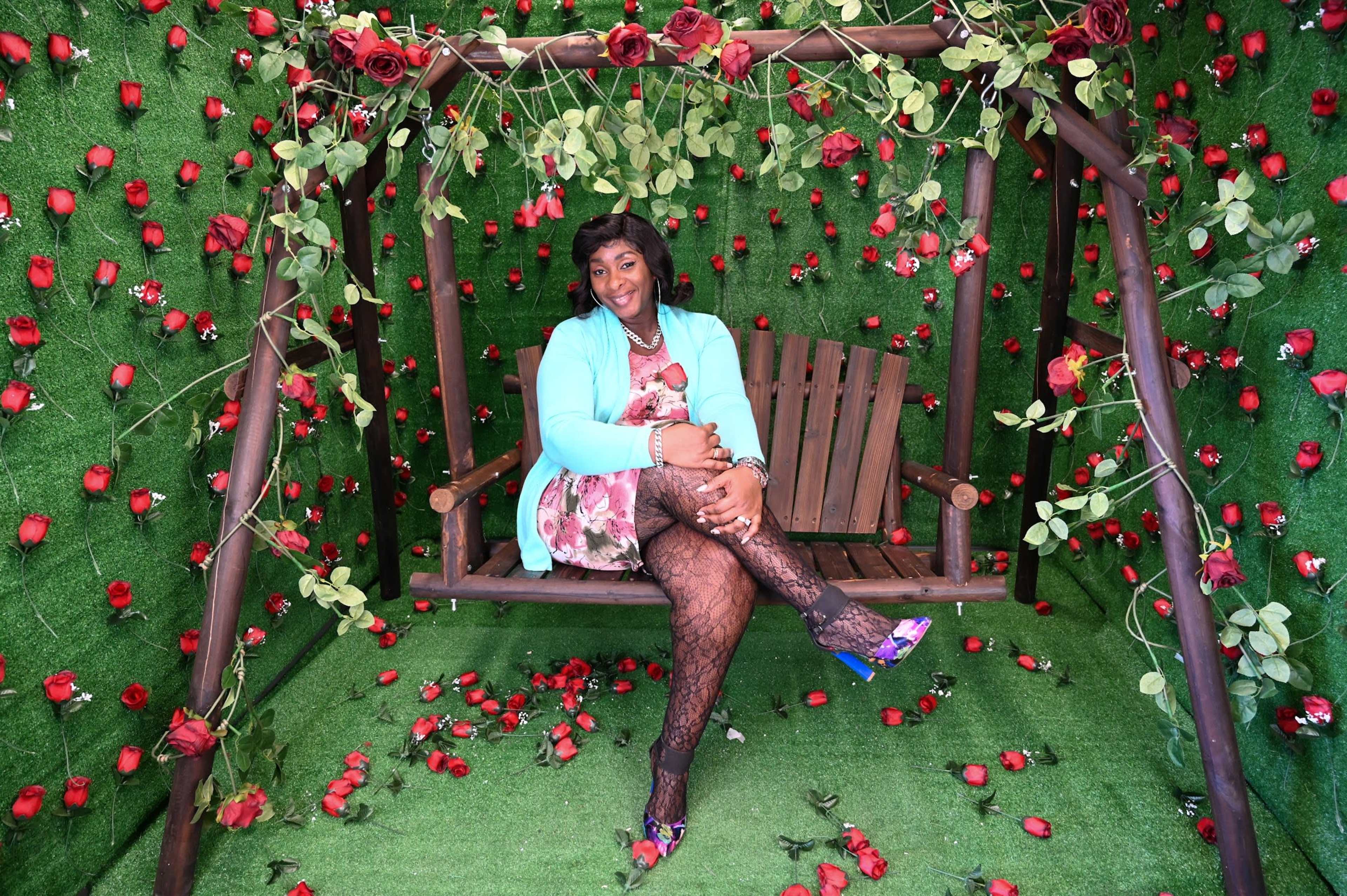 A person sits on a wooden swing surrounded by a backdrop of artificial roses and green foliage.