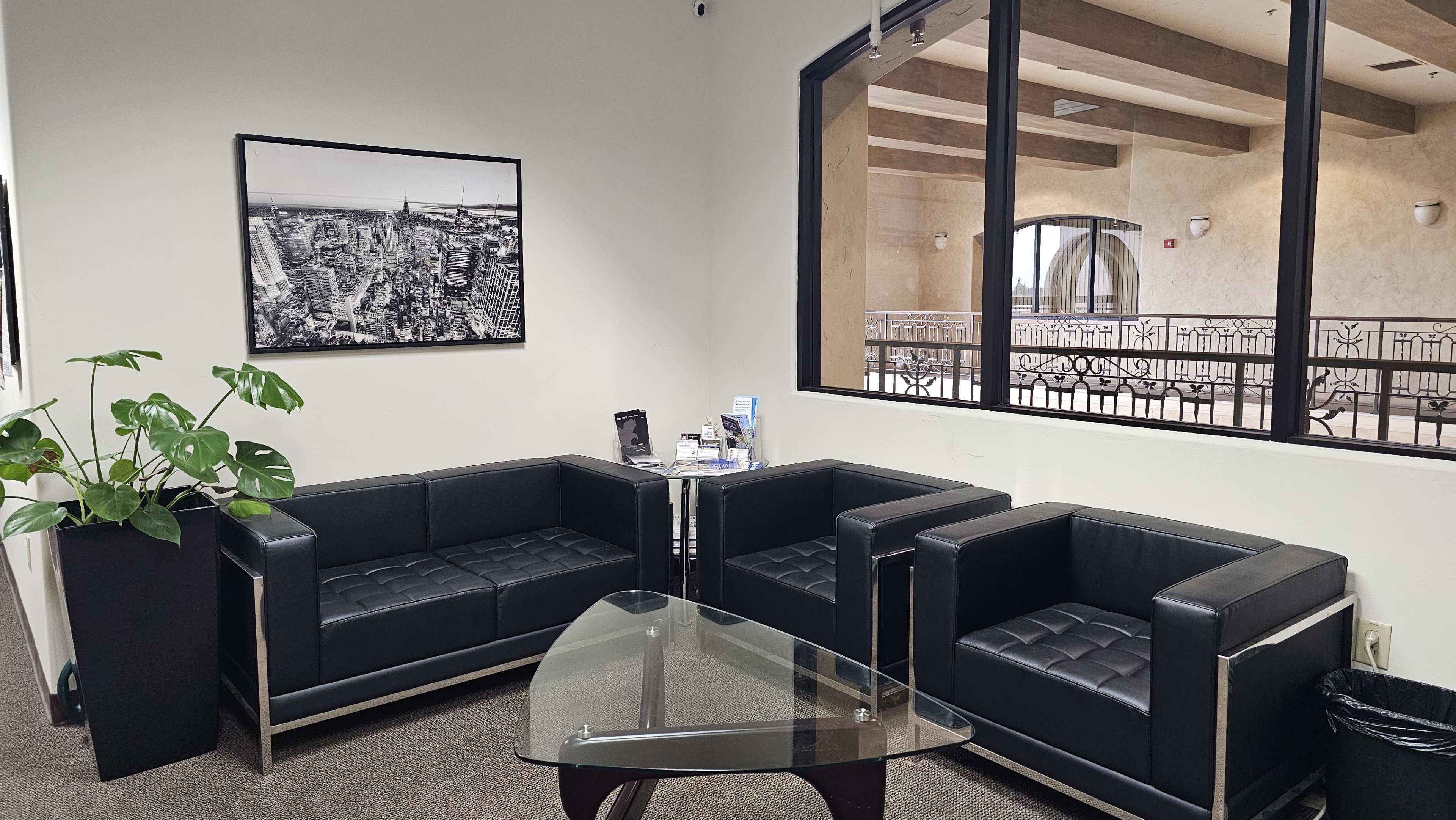 A waiting room with black leather sofas, a glass coffee table, and a large window overlooking a balcony area.