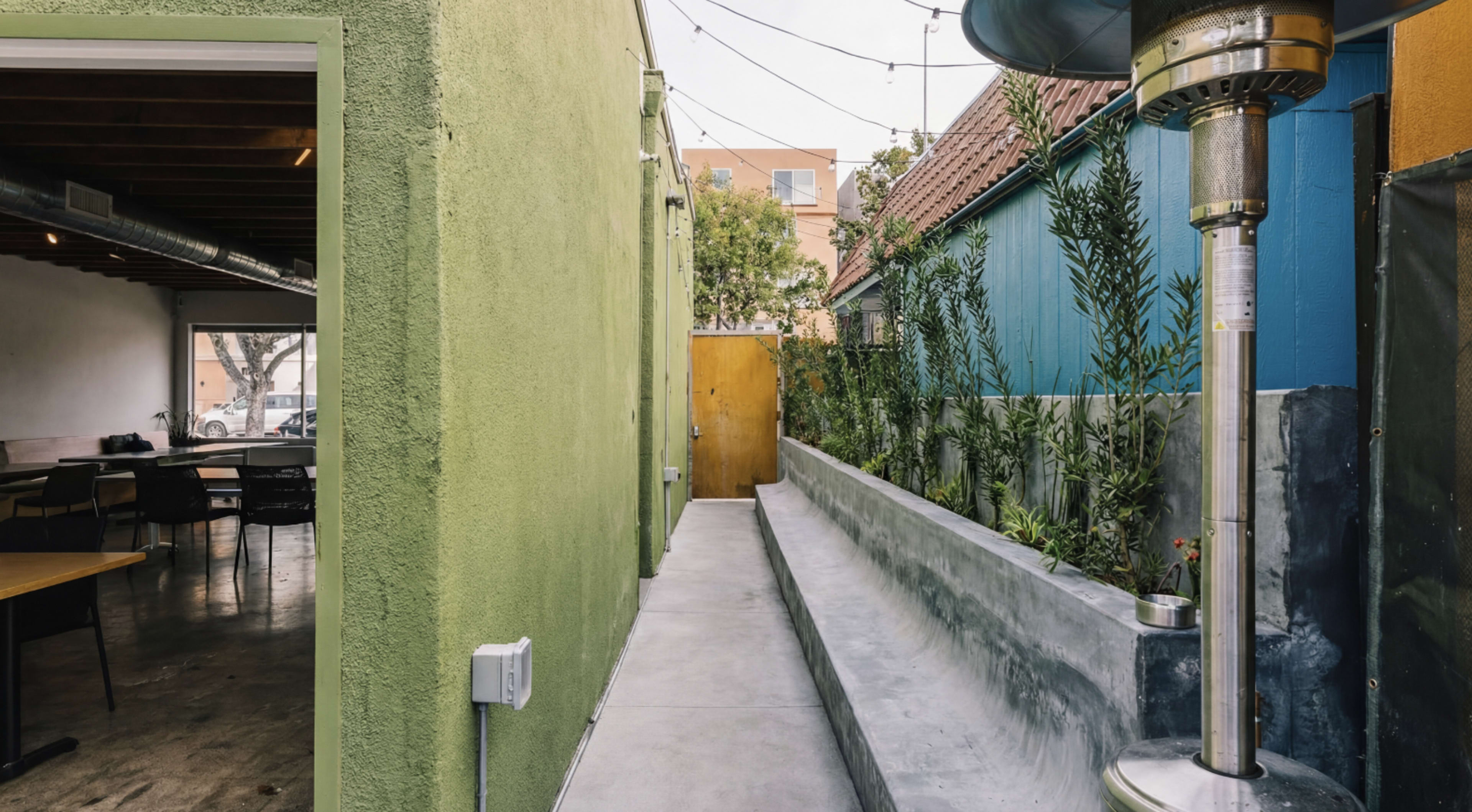 The image shows a narrow outdoor pathway lined with plants, leading to a wooden door, with a green wall on one side and a blue wall on the other.