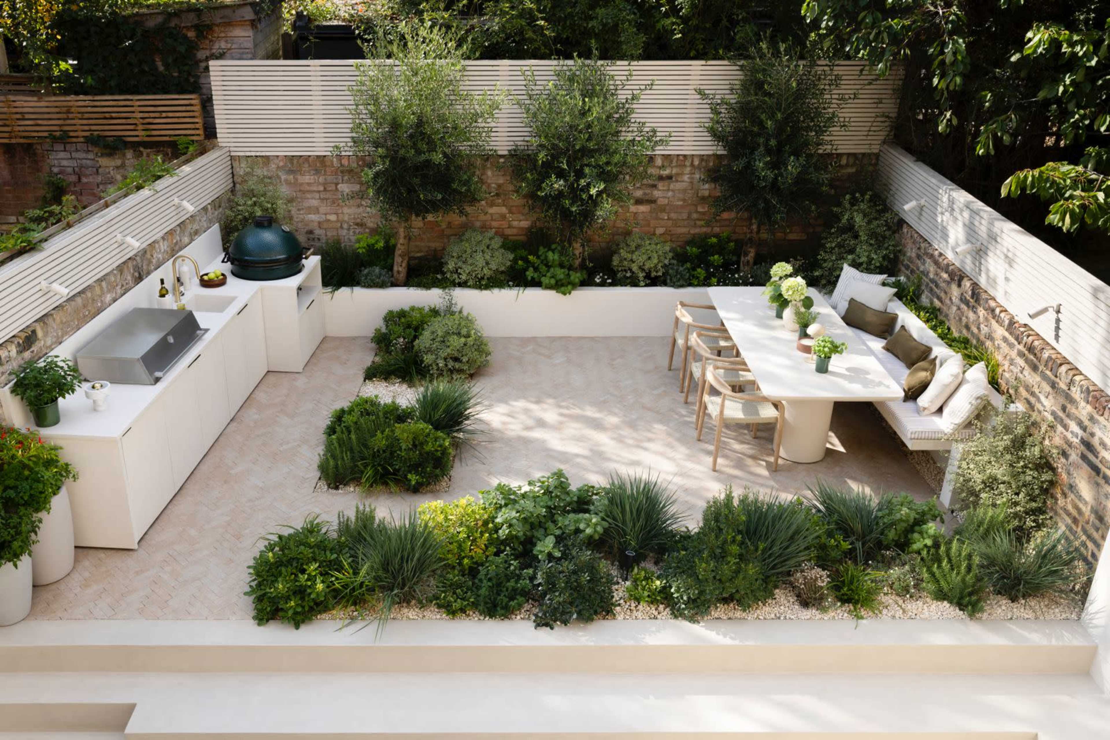 The image shows a neatly designed outdoor garden space with a seating area, a dining table, and various plants arranged around a central patio.