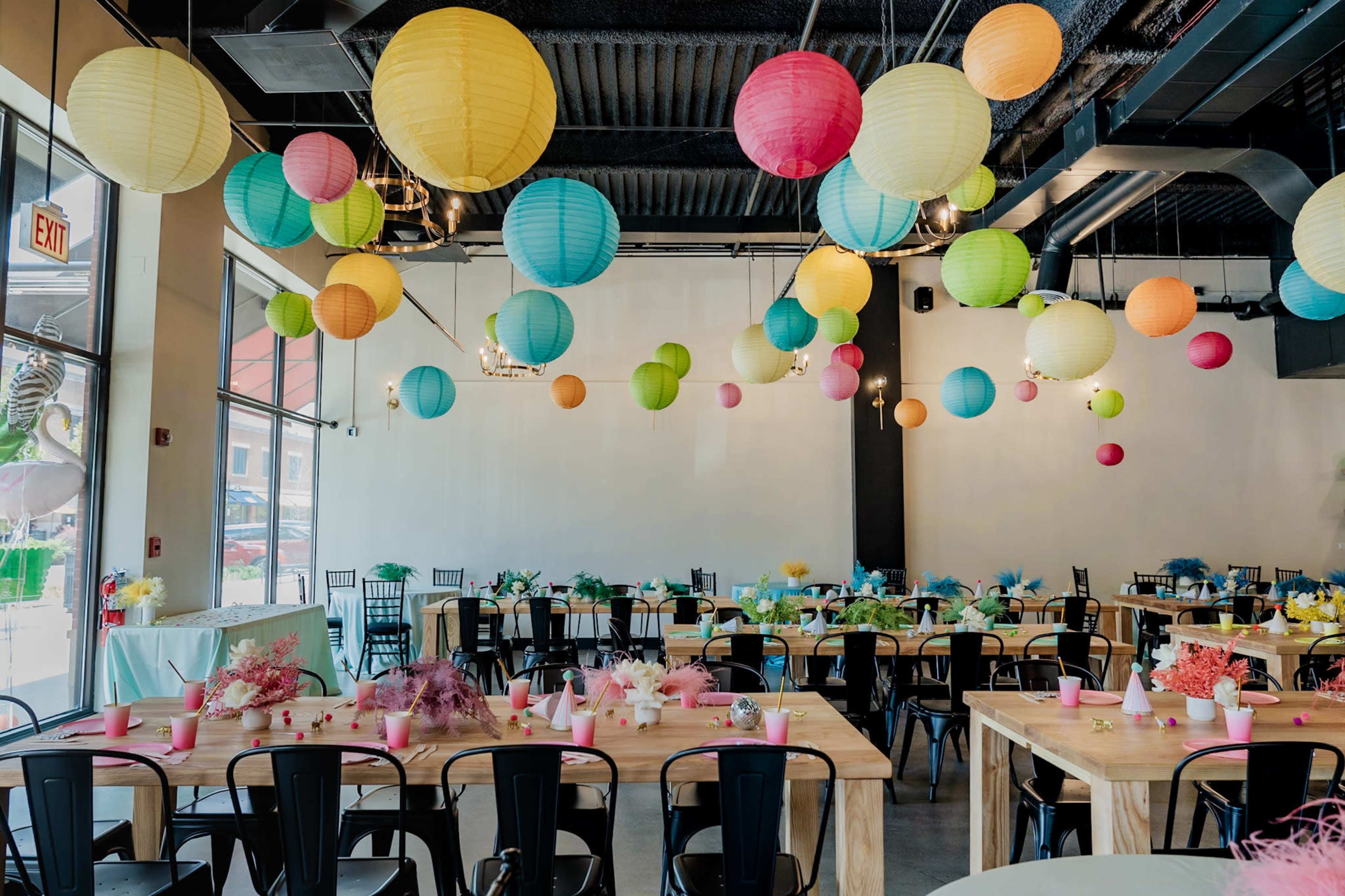A bright event space is decorated with colorful paper lanterns hanging from the ceiling above neatly arranged tables with floral centerpieces.