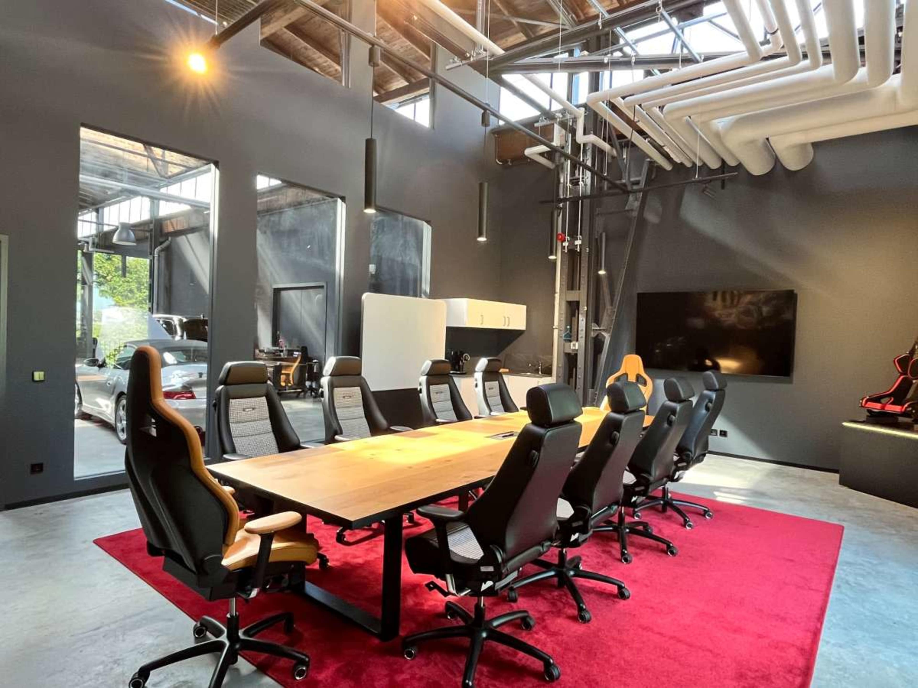 The image shows a modern conference room with a large wooden table surrounded by ergonomic chairs on a red carpet, and large windows allowing natural light to enter.