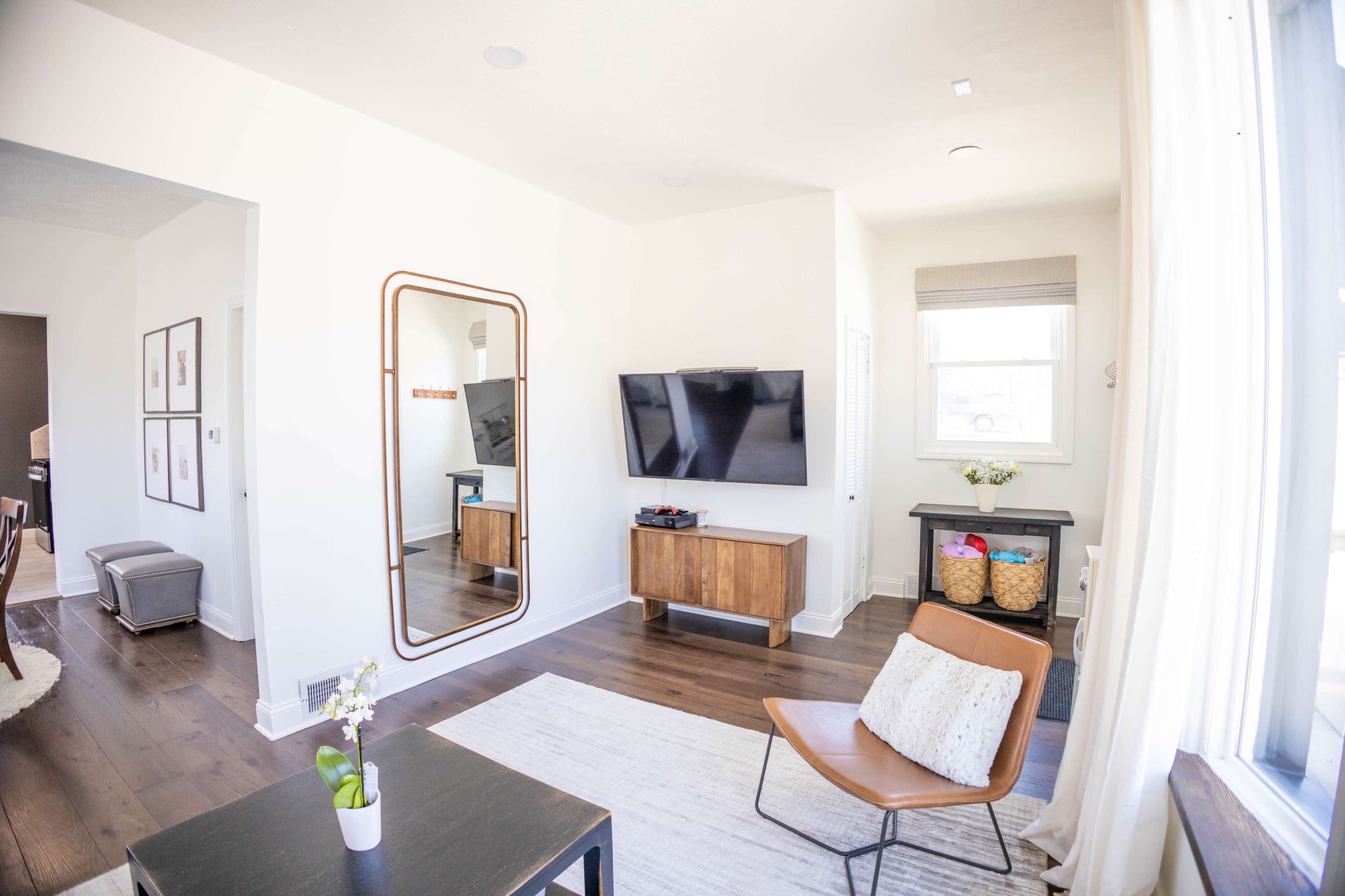 The image shows a modern living room with a large mirror, a television on a wooden console, a brown chair, and a small table.