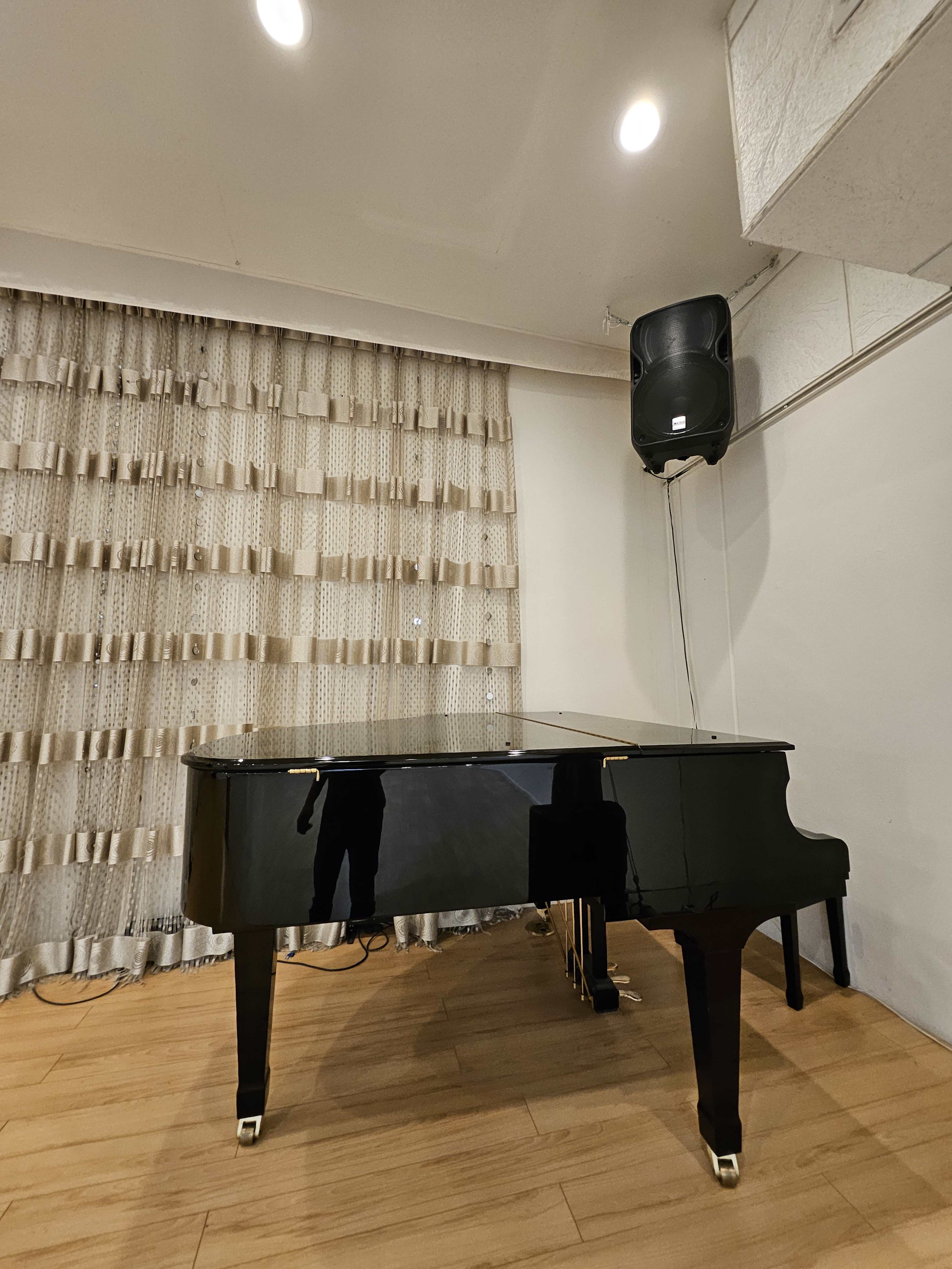 A black grand piano is positioned against a wall with textured curtains, accompanied by a mounted speaker overhead.
