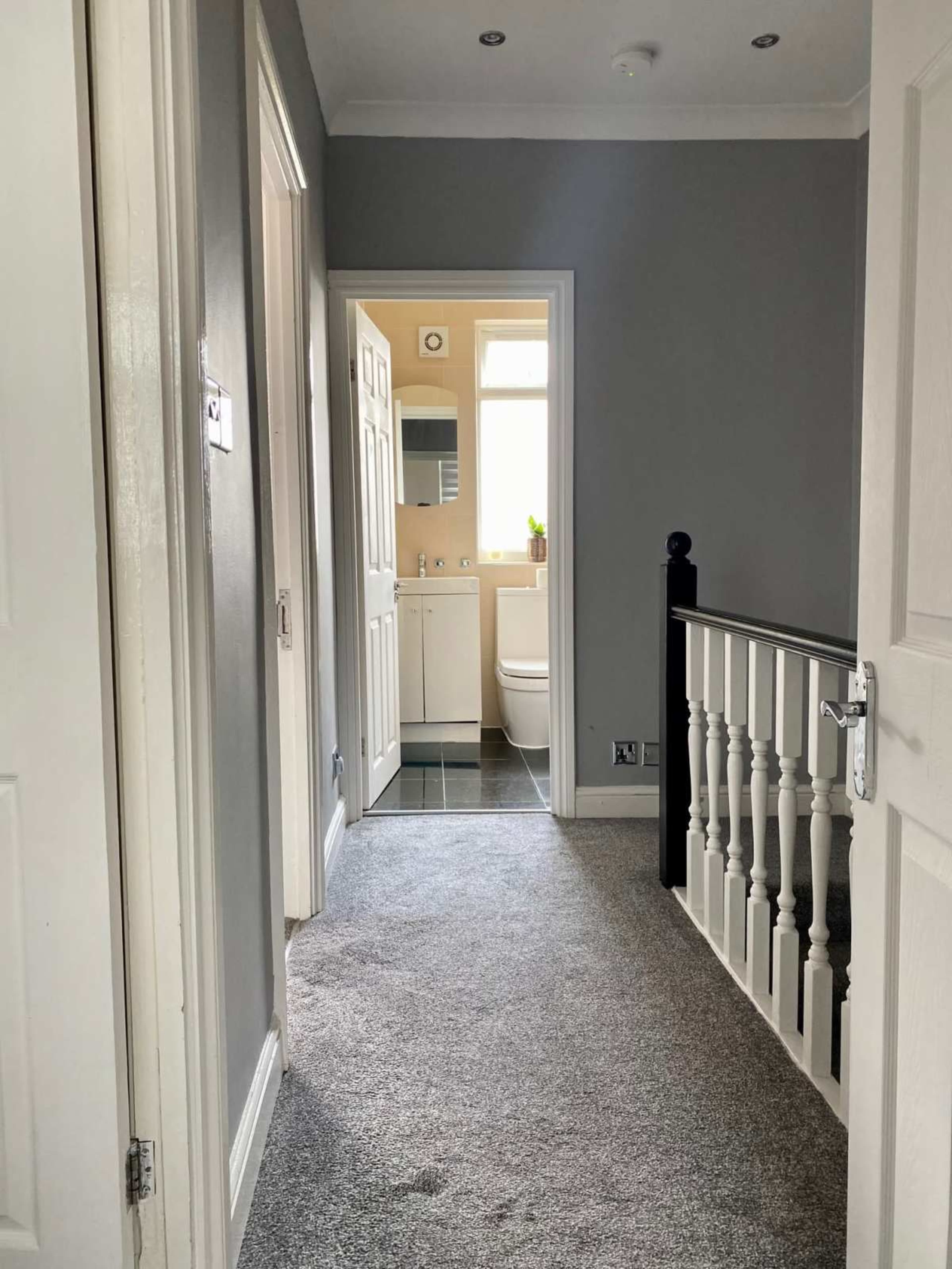 A hallway with grey walls and carpet, leading to a bathroom at the end with a small window.