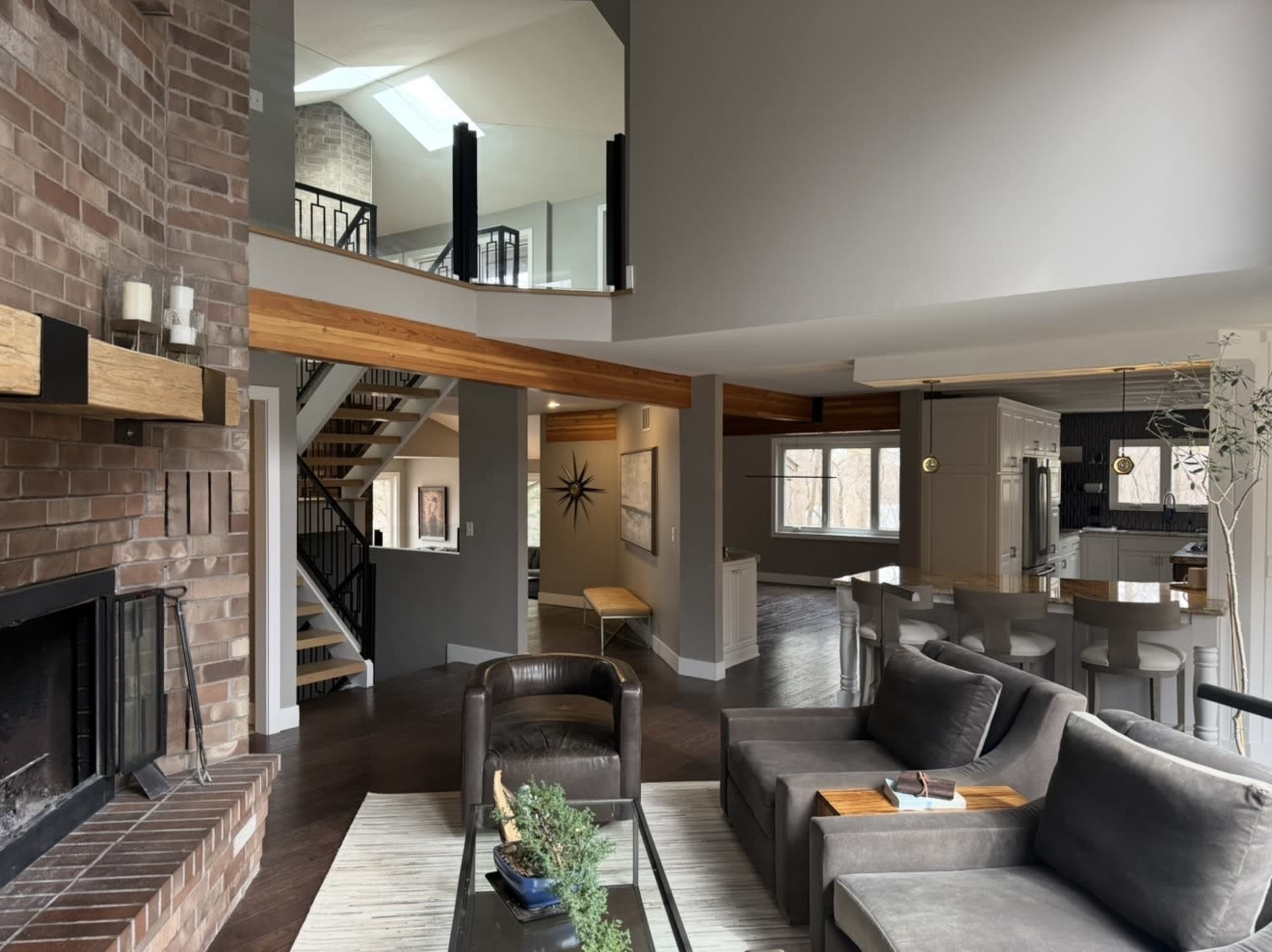 The image shows a modern open-concept living space featuring a brick fireplace, comfortable seating area, and a staircase leading to an upper level.